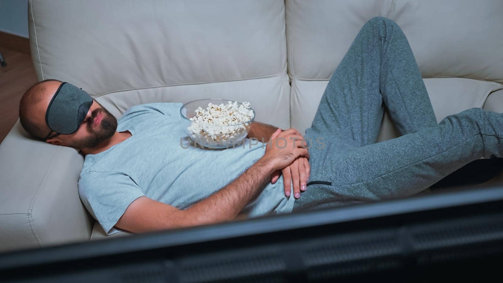 Caucasian male falling asleep while watching movie show sitting on couch late at night in kitchen. Tired man wearing eye sleep mask while the tv is on