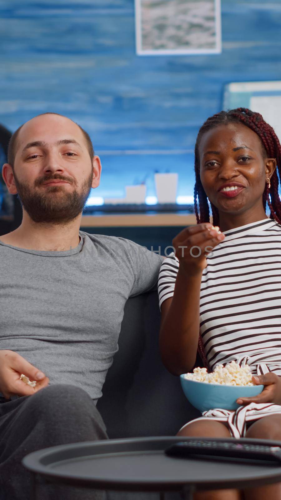 Young interracial couple watching movie on TV with popcorn sitting in living room. Mixed race partners looking at camera while eating snack and relaxing together at home on couch