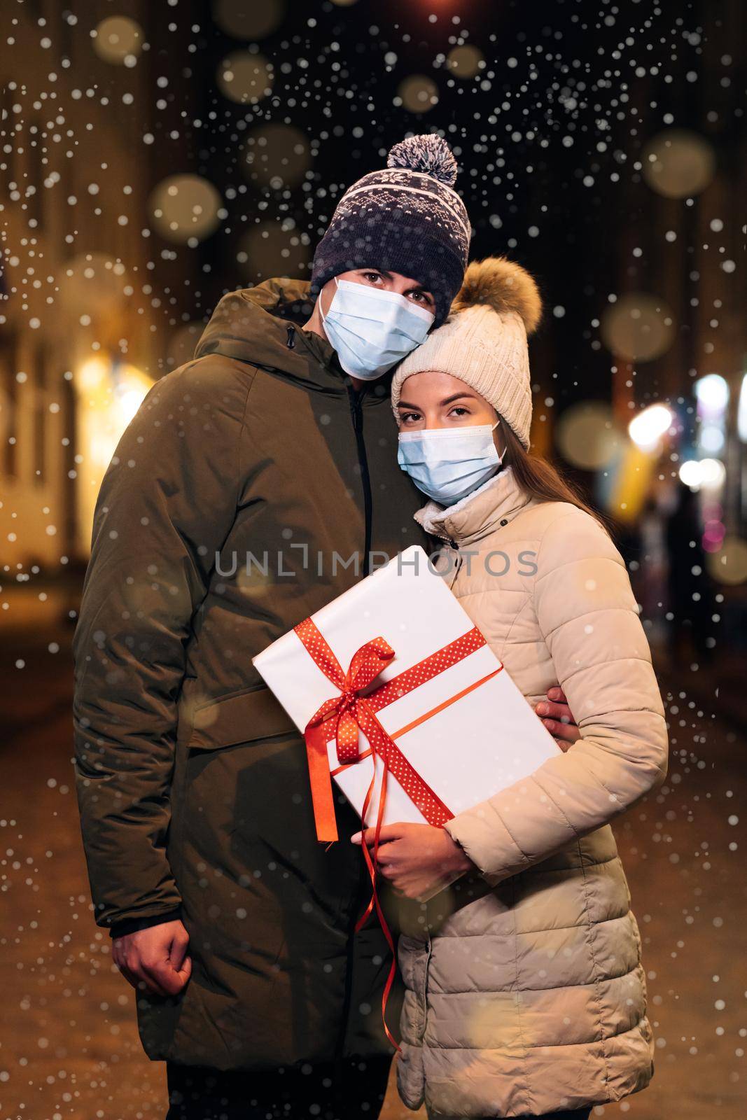 Couple in protective medical face masks celebrating xmas eve together at bright garland decoration. Christmas gifts exchange. Xmas celebration together. Lovers travelling to Europe at the height by uflypro
