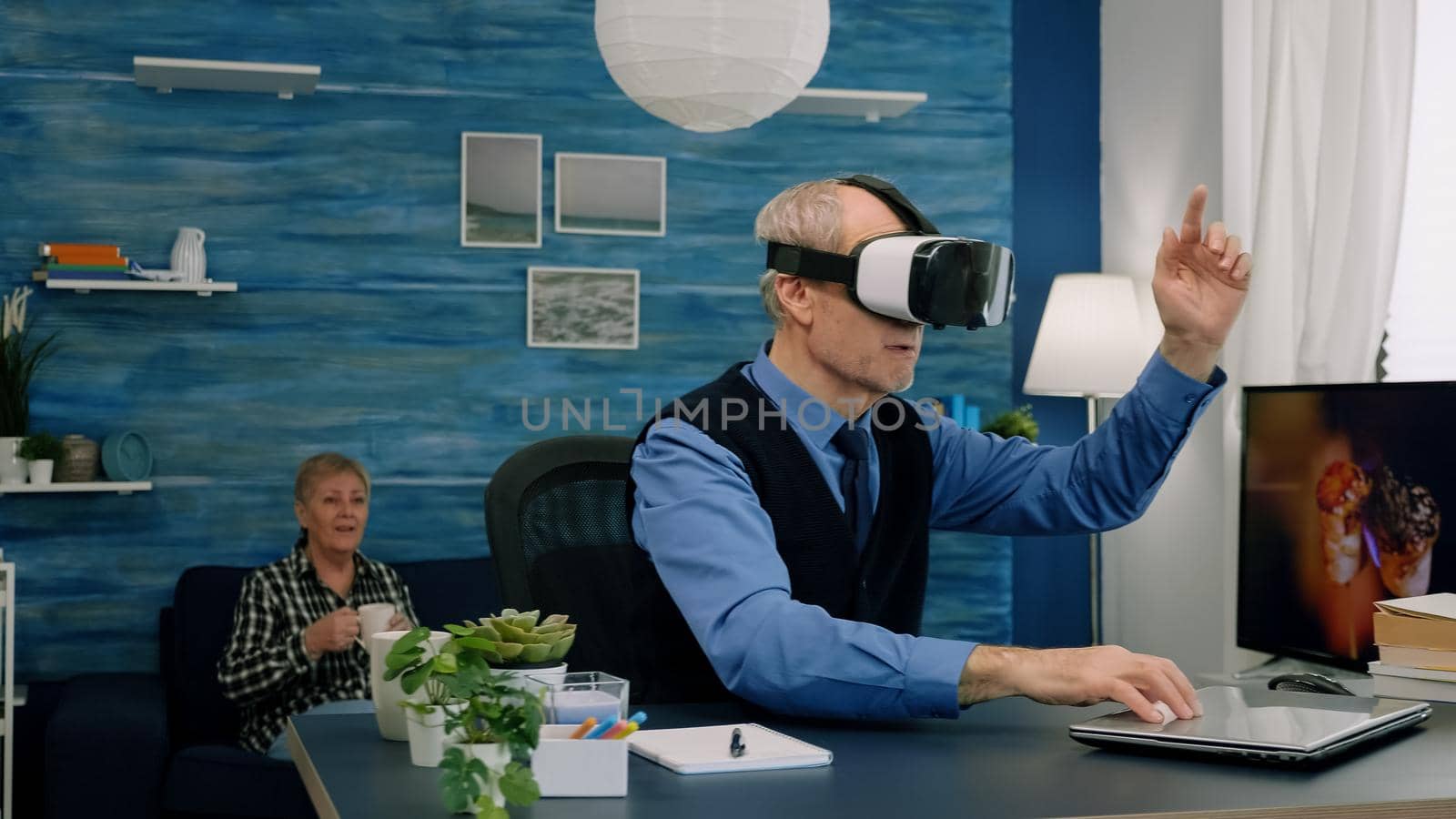 Senior man using virtual reality goggles in living room gesturing sitting in home workplace. Retired man experiencing vr headset while woman watching tv sitting on sofa in background.