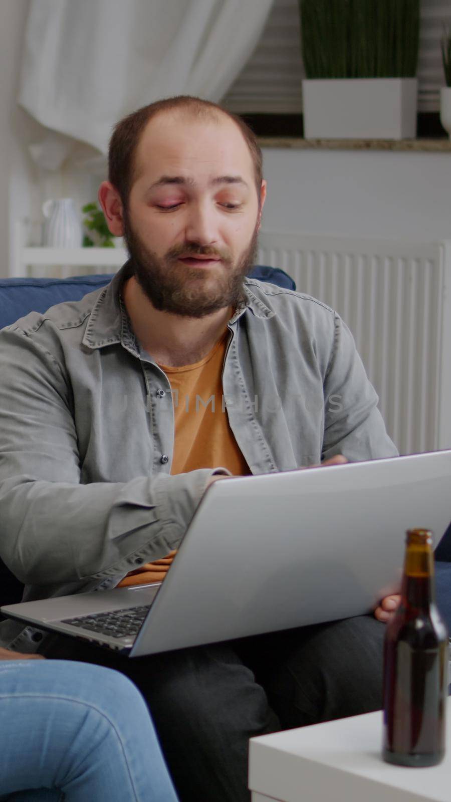 Multi-ethnic friends socializing while watching online funny videos on laptop resting on sofa. In background two womens drinking beer enjoying time spend together during entertainment party