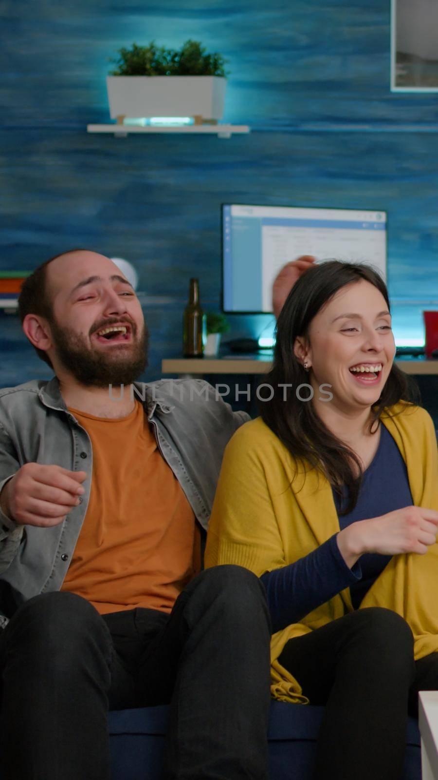 Multiracial friends laughing smiling having fun together while sitting on couch in front of TV watching comedy movie. Group of mixed race people enjoying free time drinking beer eating snacks