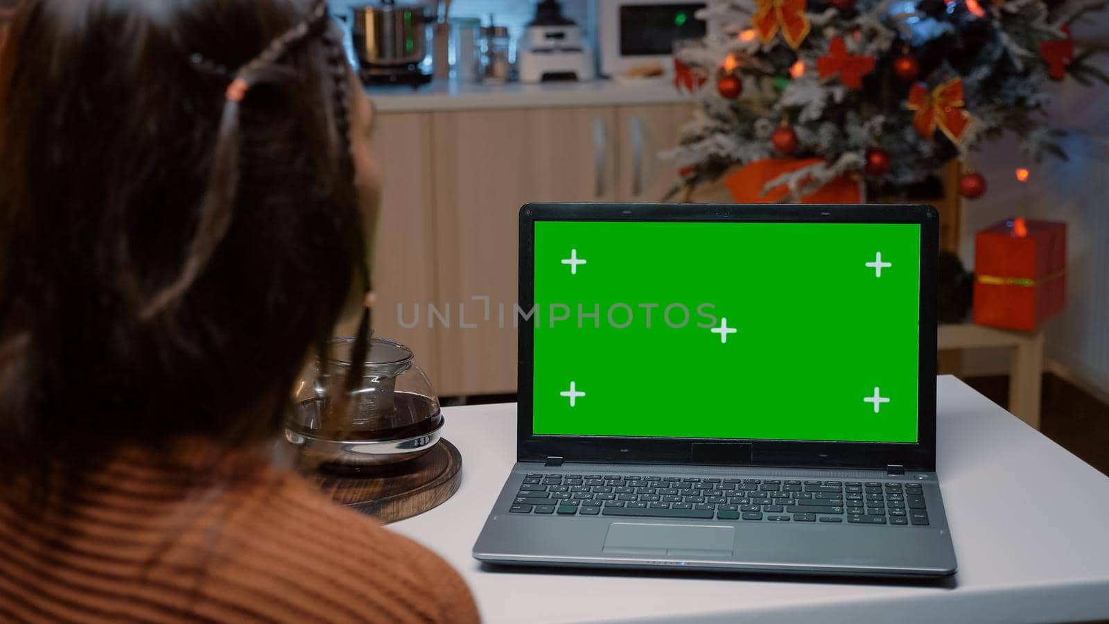 Caucasian woman looking at green screen on laptop while sitting in festive kitchen at home with winter decorations. Young adult using modern device preparing for christmas dinner party