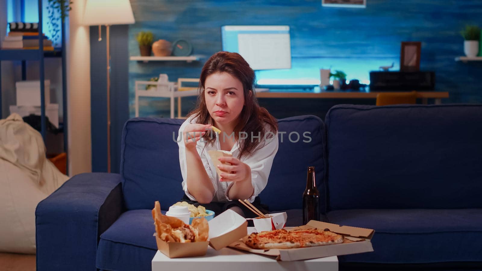 Young adult using video call remote technology sitting on living room sofa. Woman talking on online conference while eating fries and fast food. Person with internet and takeaway food