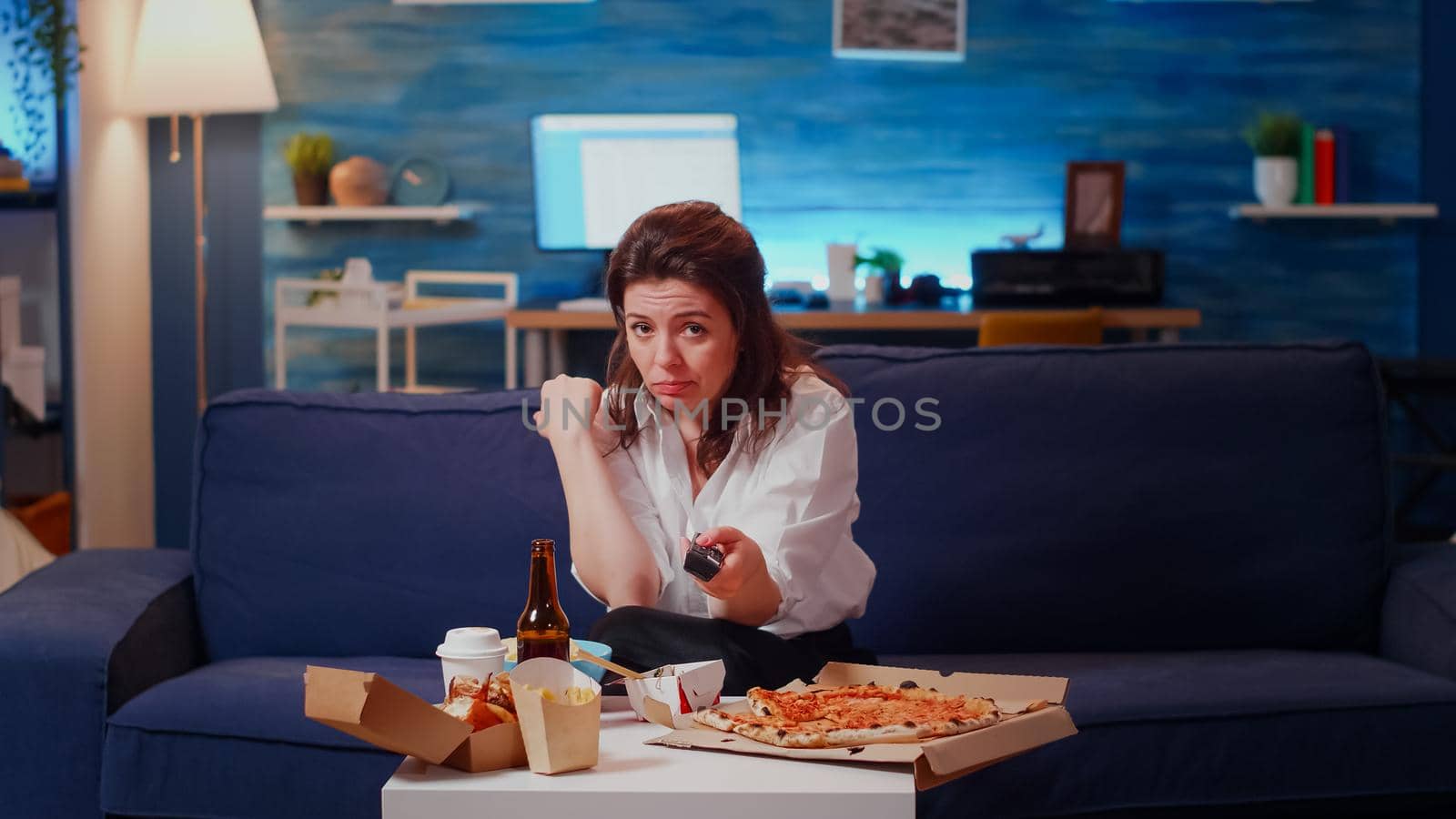 POV of woman switching channels and eating chips on living room couch. Young adult looking at camera watching television and enjoying snack or takeaway meal with fast food and beer