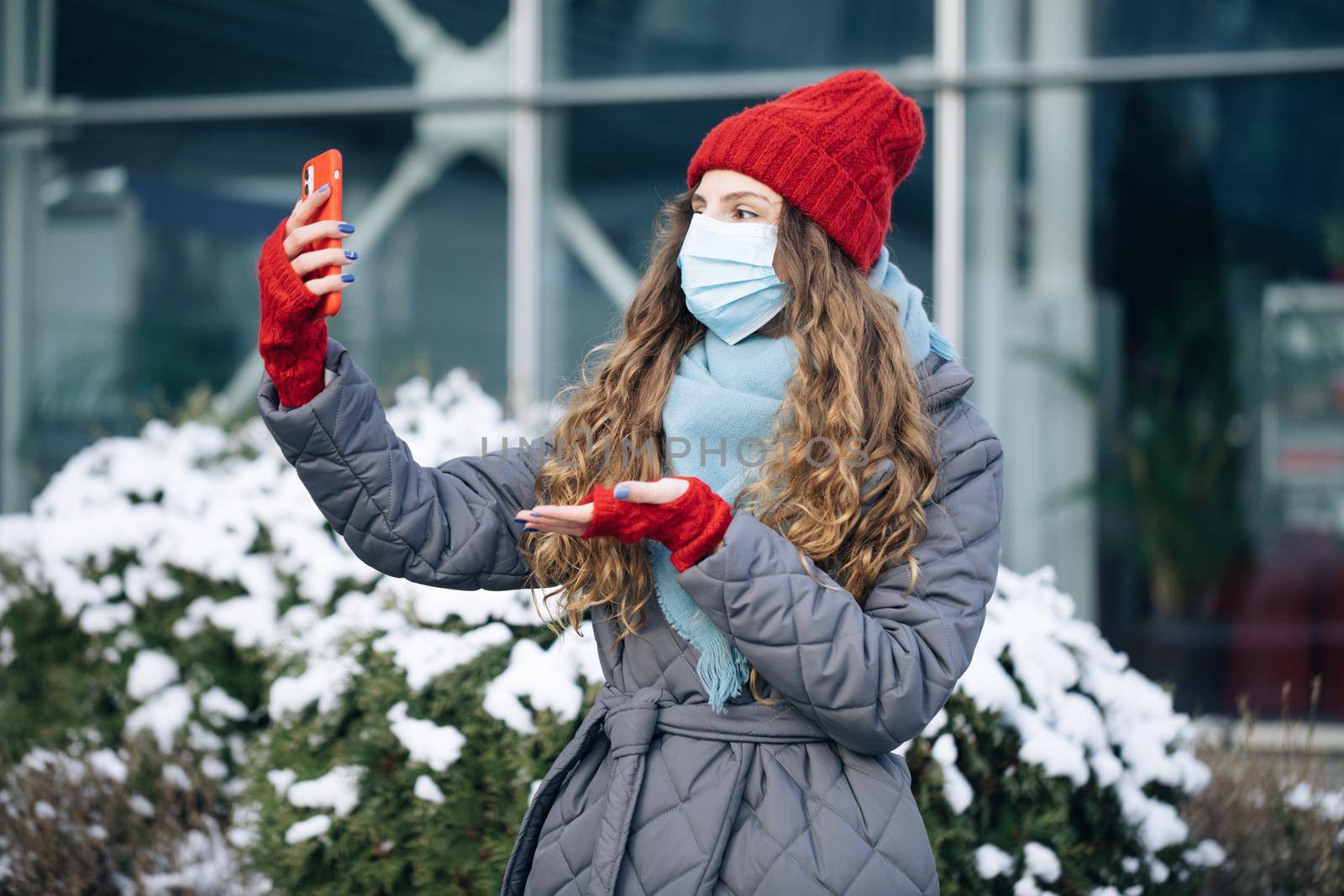 Female blogger in face mask is making video talking for her social page at the street in winter time. Woman look around on the winter street city. Being online, social networks by uflypro