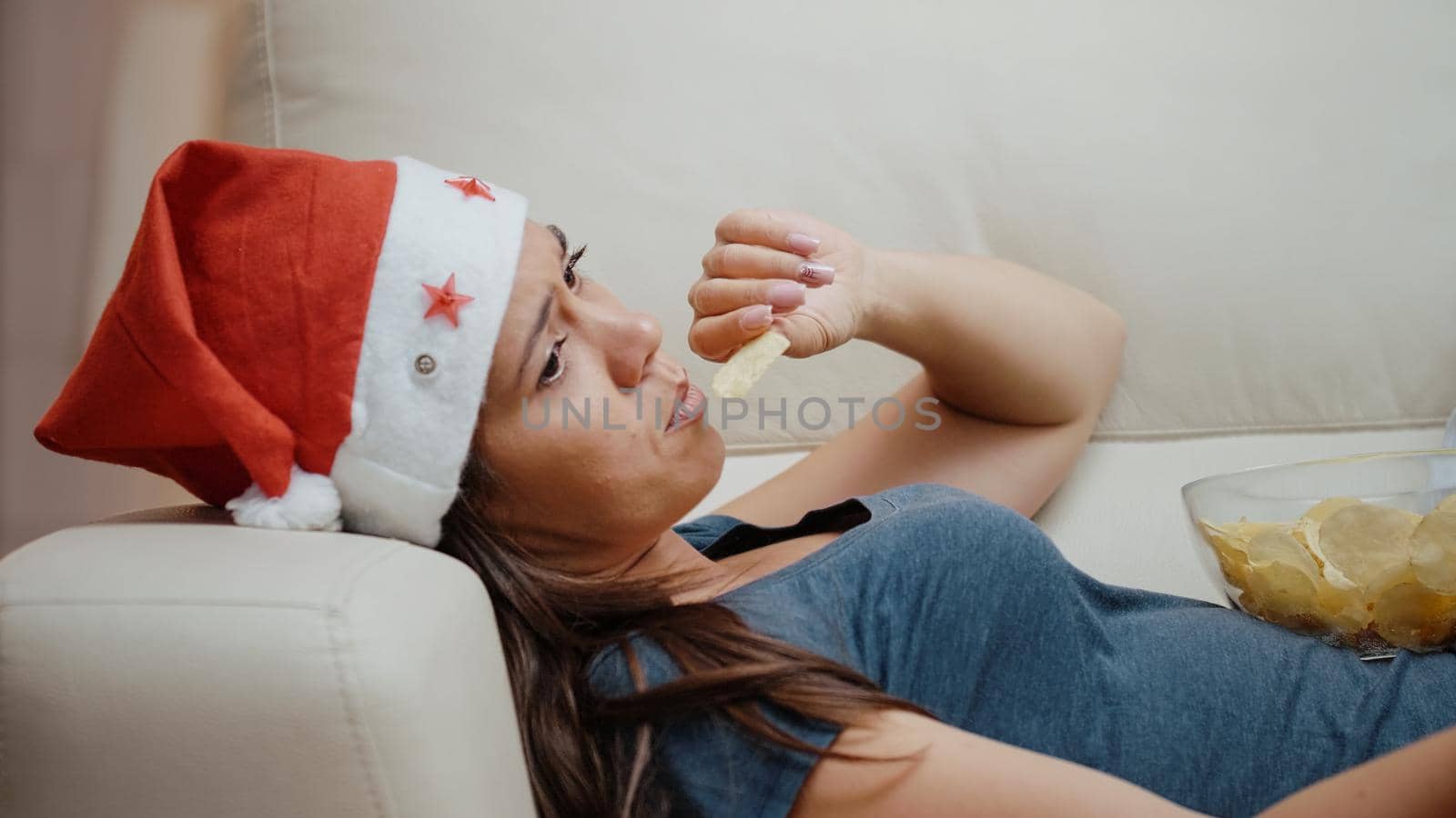 Bored woman wearing santa hat and eating chips from bowl while watching television and using TV remote control to switch channels on couch to celebrate christmas holiday. Person eating