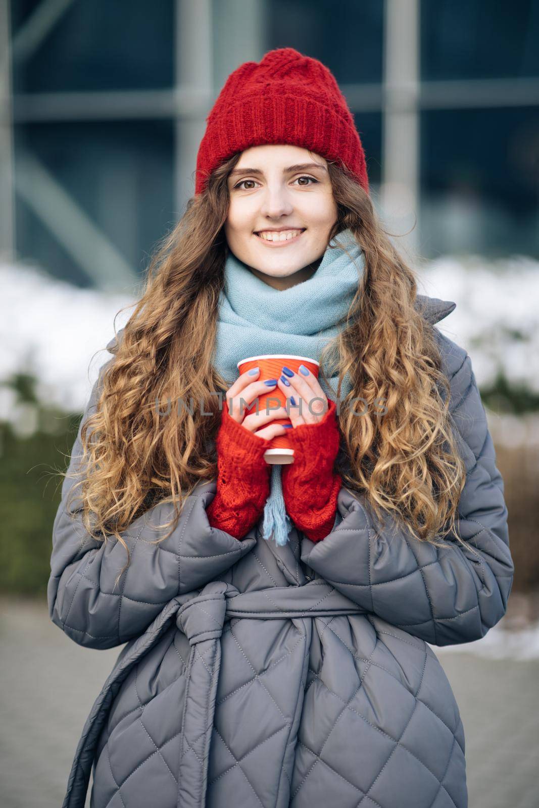 Girl in a red warm hat with a cup of coffee in her hands stands on the street in winter. Portrait of nice-looking curly caucasian elegant young woman looking into camera. Female portraits.