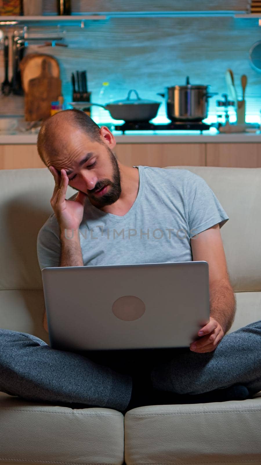Tired man in pajams browsing on internet lifestyle information using laptop computer sitting on sofa. Freelancer working at online project in front on television late at night in kitchen