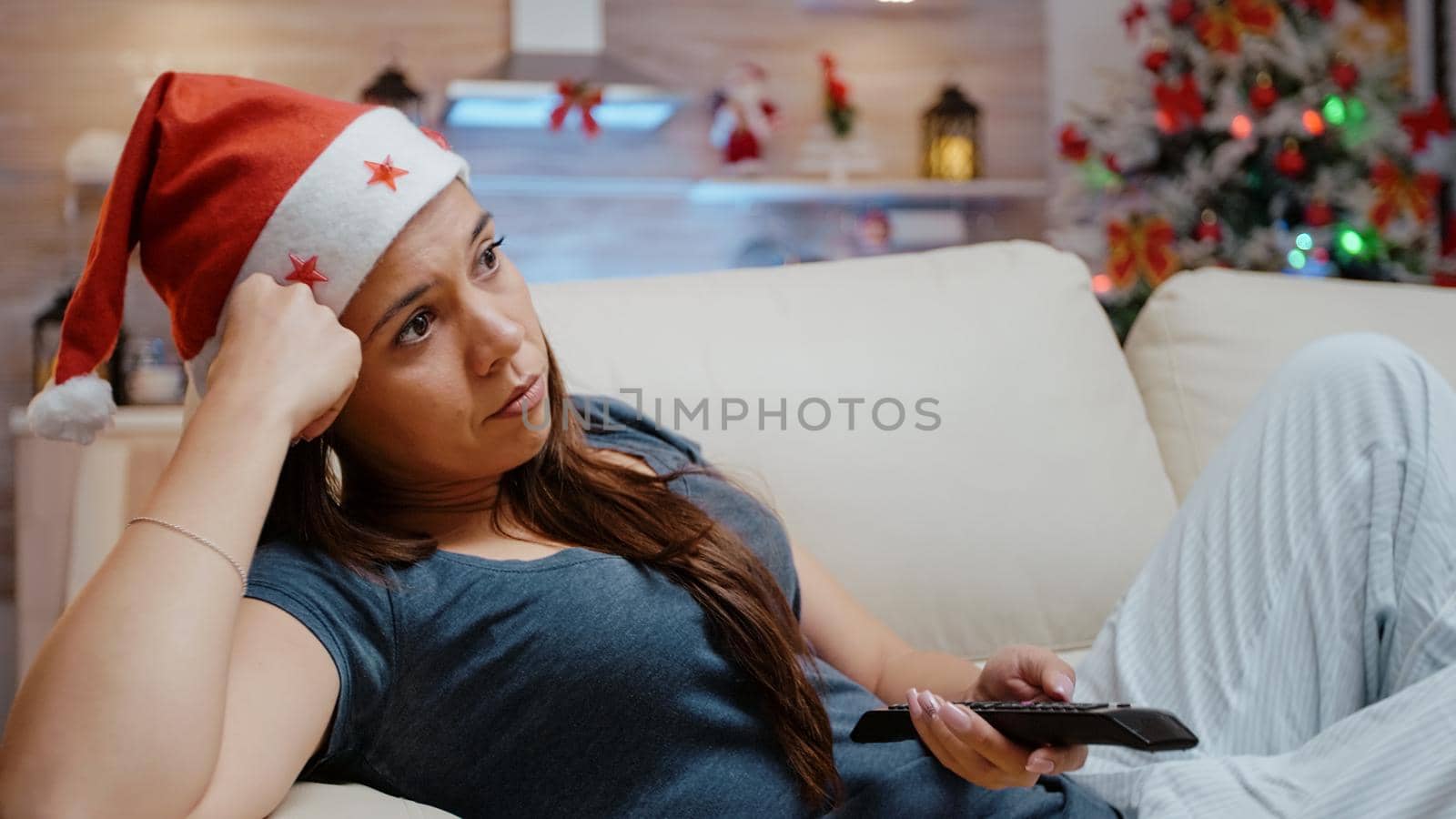 Woman switching channels with TV remote control feeling bored on christmas eve holiday. Person wearing santa hat while laying on sofa and looking at movie on television. Festive adult