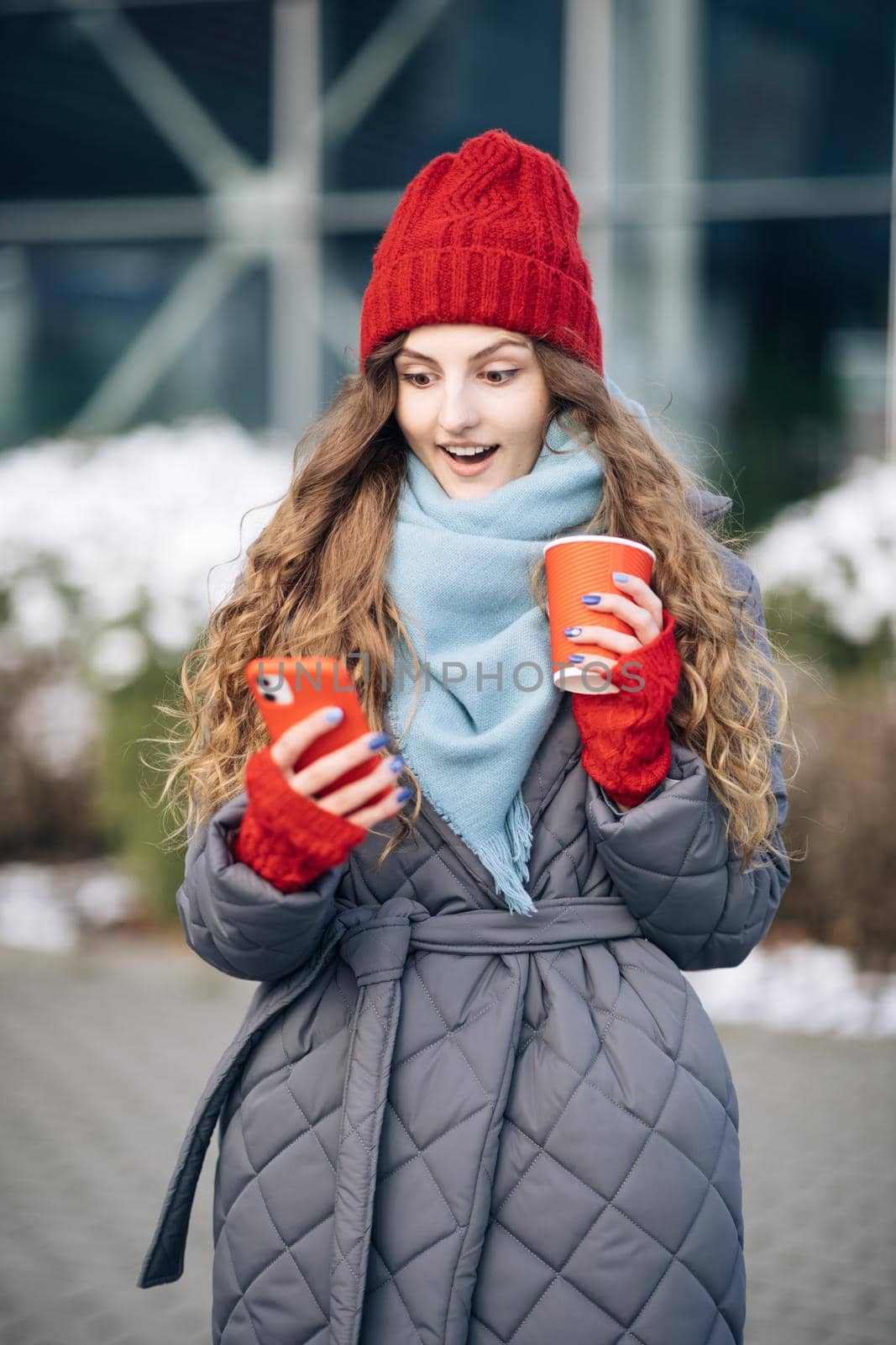 Portrait of young beautiful curly woman in winter clothes holding phone celebrate good mobile news surprise bid. Excited woman winner screaming yes rejoicing success looking at cellphone by uflypro