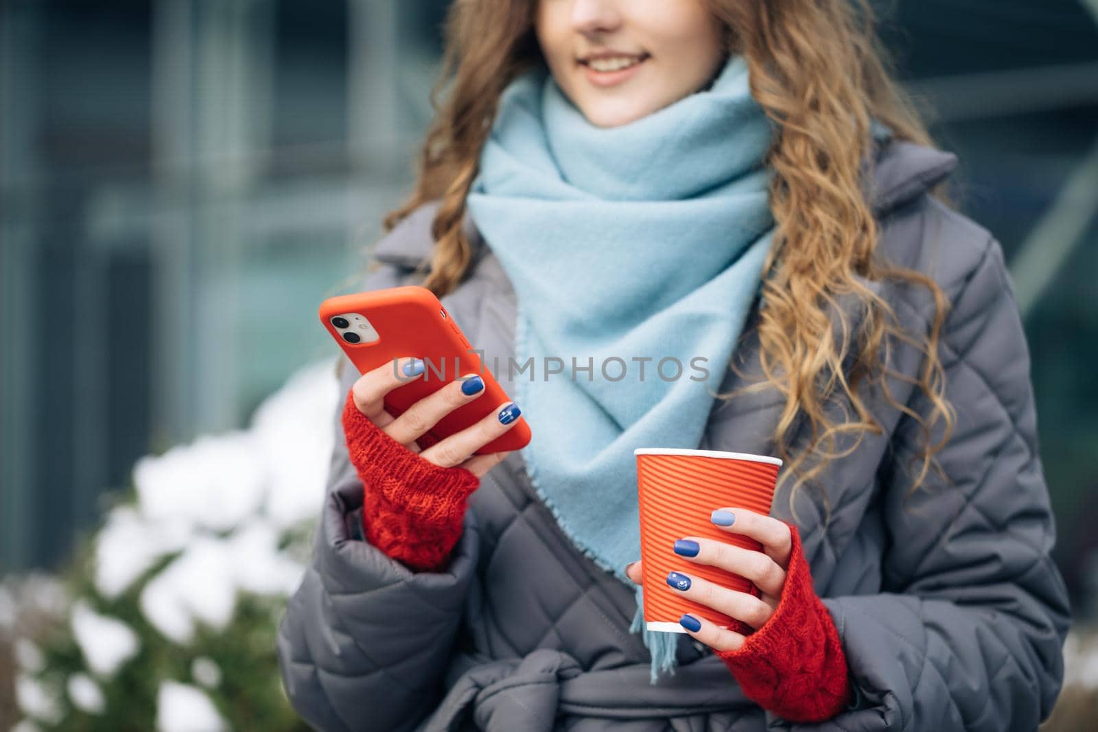 Curly-haired Young Girl is Laughing and Reading Something on Her Smartphone While Holding a Cup of Coffee in Other Hand. Woman Walking look around on the street city. Being online, social networks.