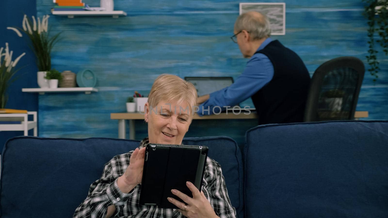 Senior woman reading emails on digital tablet sitting on sofa at home, while senior adult man working at laptop in background. Person enjoying using notepad browsing internet shopping