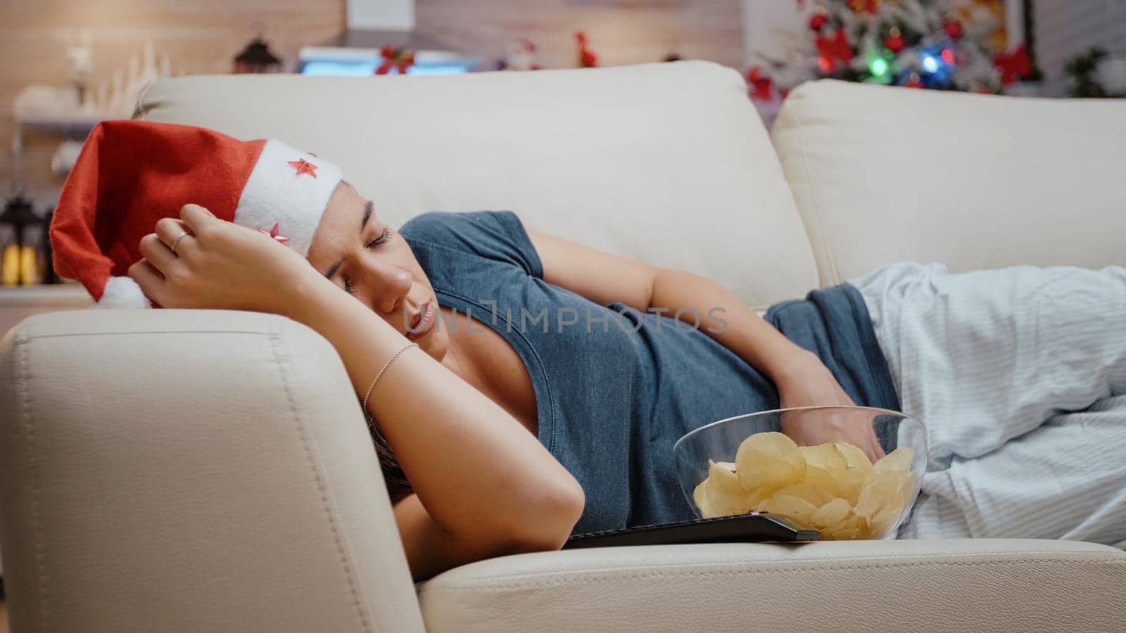 Close up of adult sleeping on couch with bowl of chips and TV remote control. Woman wearing santa hat resting on christmas holiday at television screen. Person alseep on sofa.