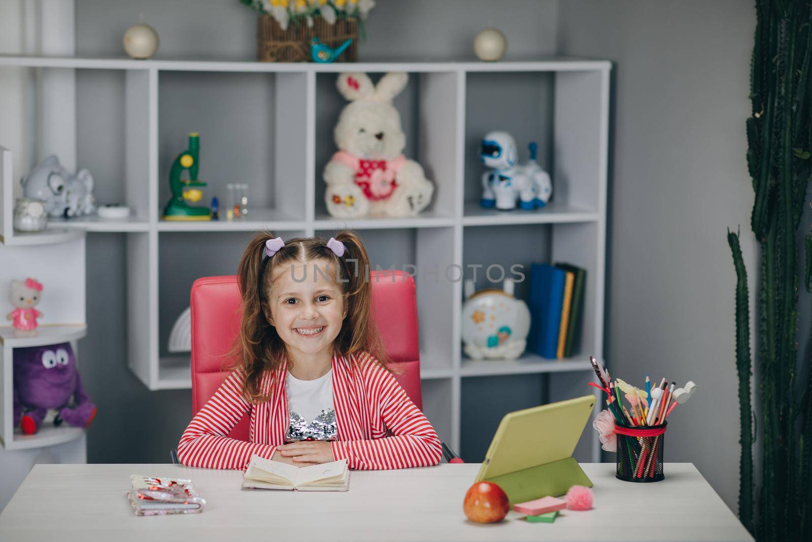 Portrait of a preschool girl looking at the camera and smiling. Kid girl sitting at the desk in children room at home and studying online. Distance learning, school online. by uflypro
