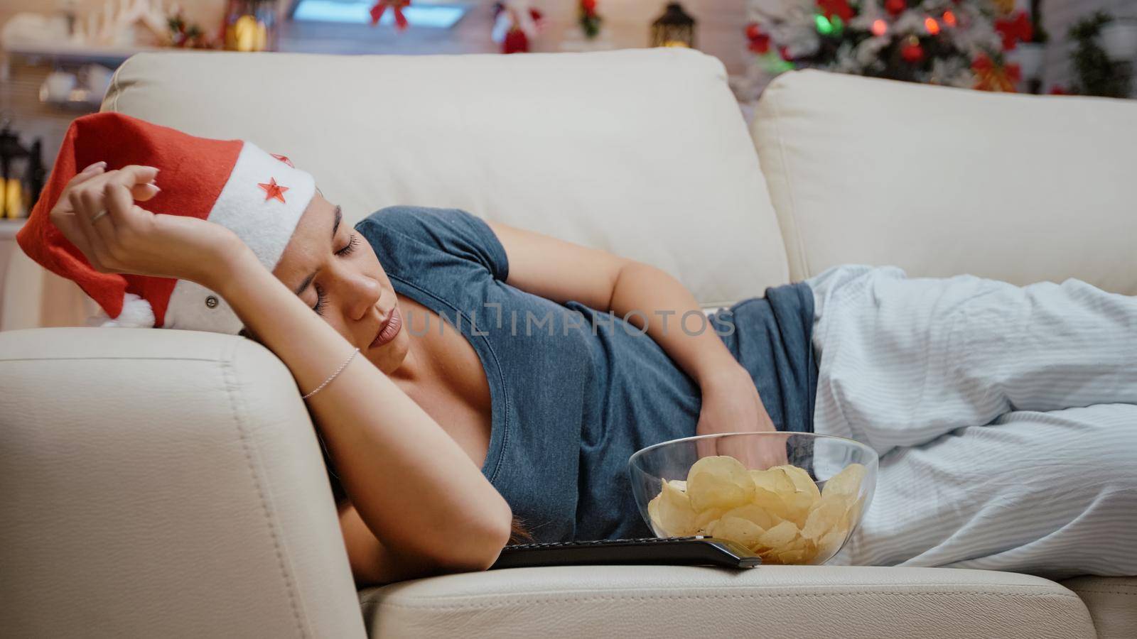 Close up of woman falling asleep while watching television on couch. Festive adult feeling sleepy, looking at TV on christmas eve celebration day. Tired person wearing santa hat