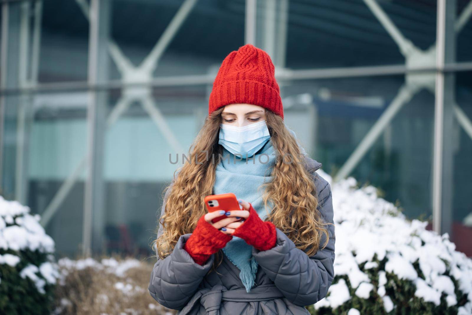 Portrait of Caucasian curly female in mask browsing on smartphone while standing on street. Joyful young woman texting on cellphone. Curly female standing on winter street by uflypro
