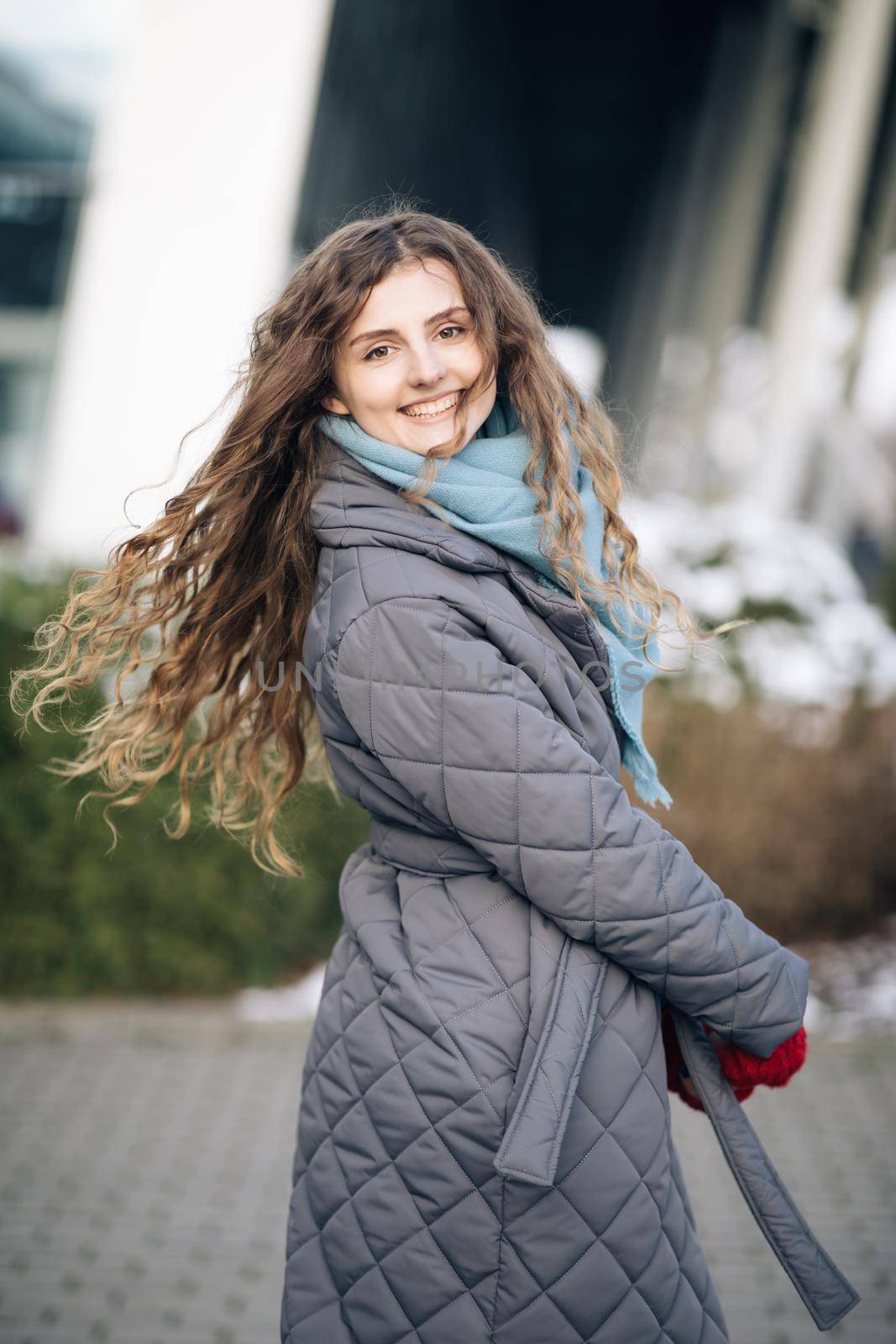 Charming young curly woman with a magnificent brown hair, big brown eyes and stylish look turns to camera and smiles. Attractive young lady is rushing in the winter city-center