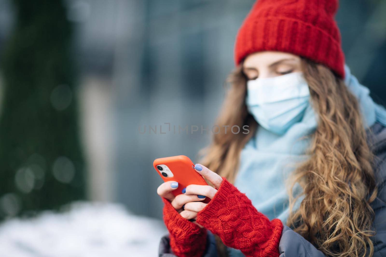 Close up portrait of beautiful Caucasian young curly hair female in medical mask texting on cellphone. Caucasian woman browsing on smartphone and looking away. Being online, social networks