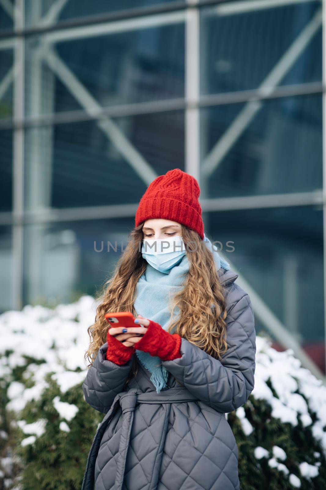 Cheerful Caucasian beautiful curly female in mask browsing on smartphone while standing on street. Joyful young woman texting on cellphone. Curly female standing on winter street by uflypro