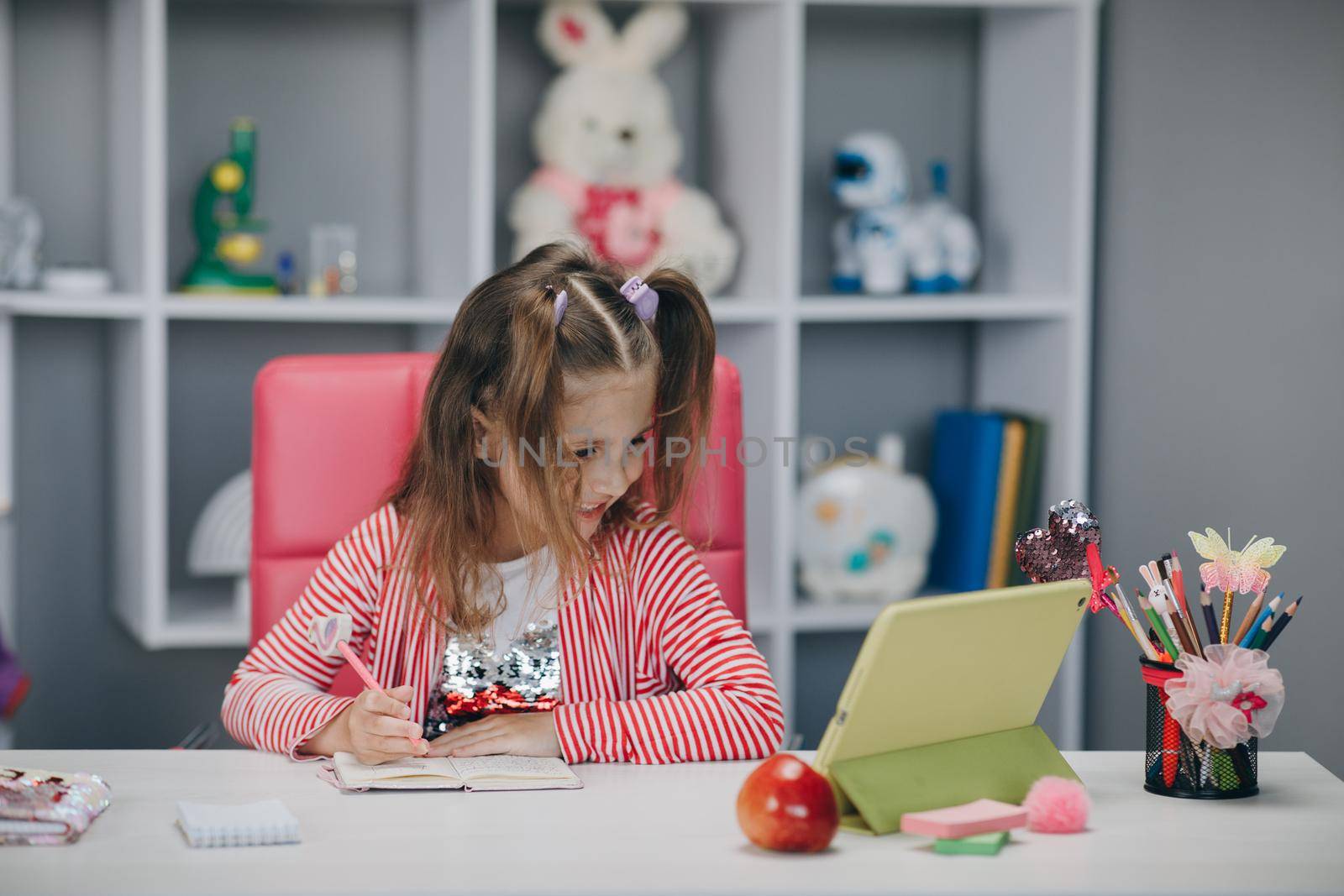 Young preschool girl watching lesson online and studying from home. Kid girl taking notes while looking at computer screen following professor doing math on video call