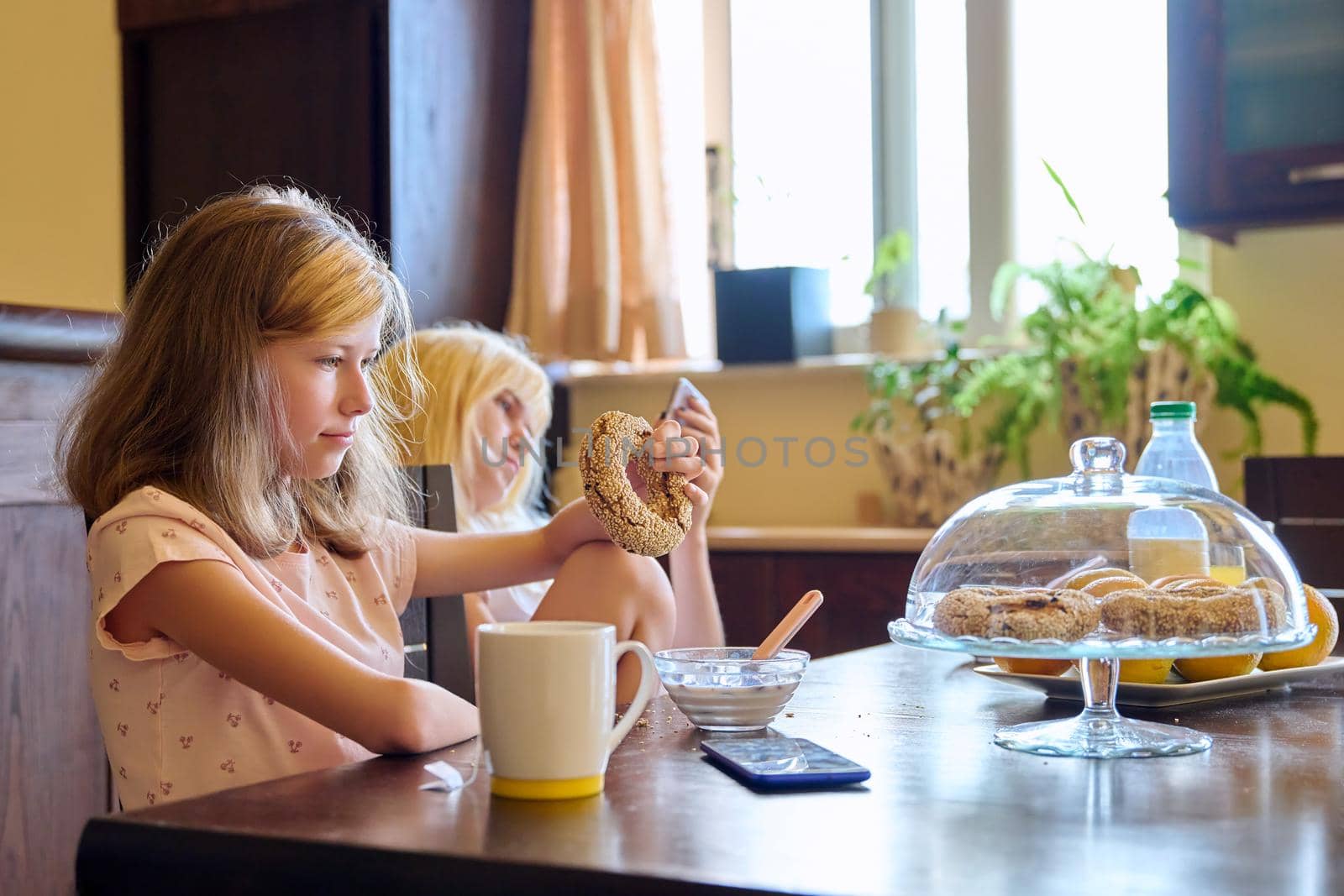 Family, children, two girls, sisters eating at home. Teenagers having breakfast in kitchen, milk fruit snacks on table, older girl with smartphone. Adolescence, home everyday life, real life, lifestyle