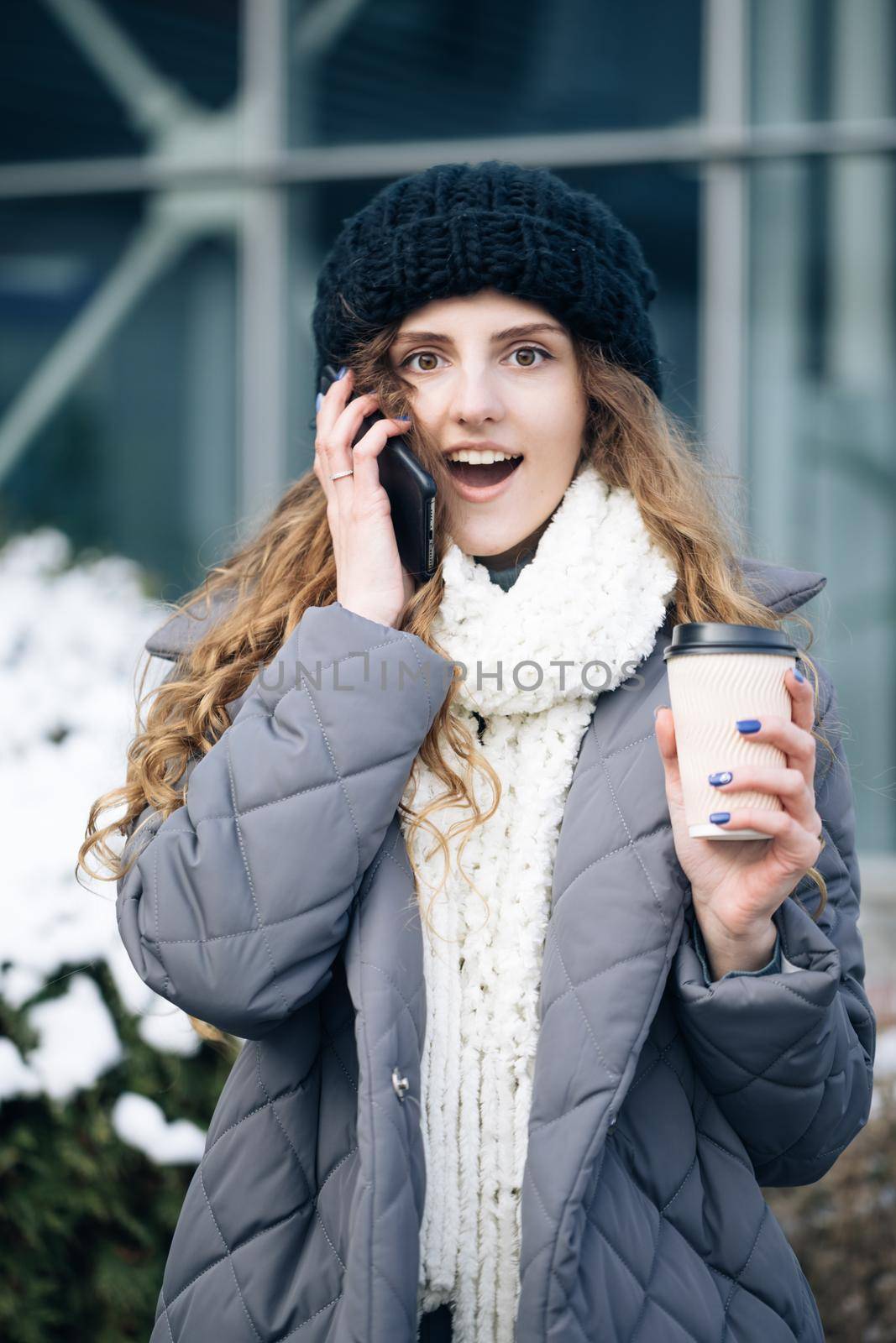 Young girl talking on her mobile phone while walking in the city street. Woman using her smartphone and holding paper cup with coffee.