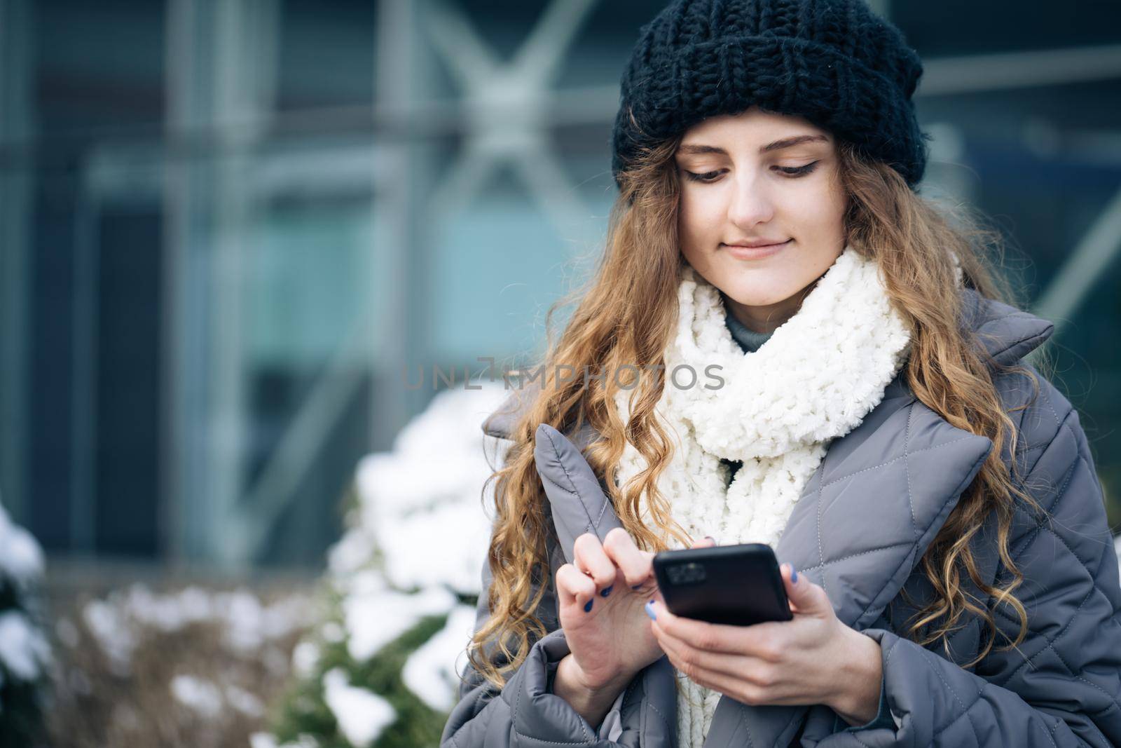 Female using smartphone standing outside. Smiling happy girl employee typing on a cellphone. Joyful young female in good mood typing and scrolling on smartphone outdoors. Winter concept by uflypro