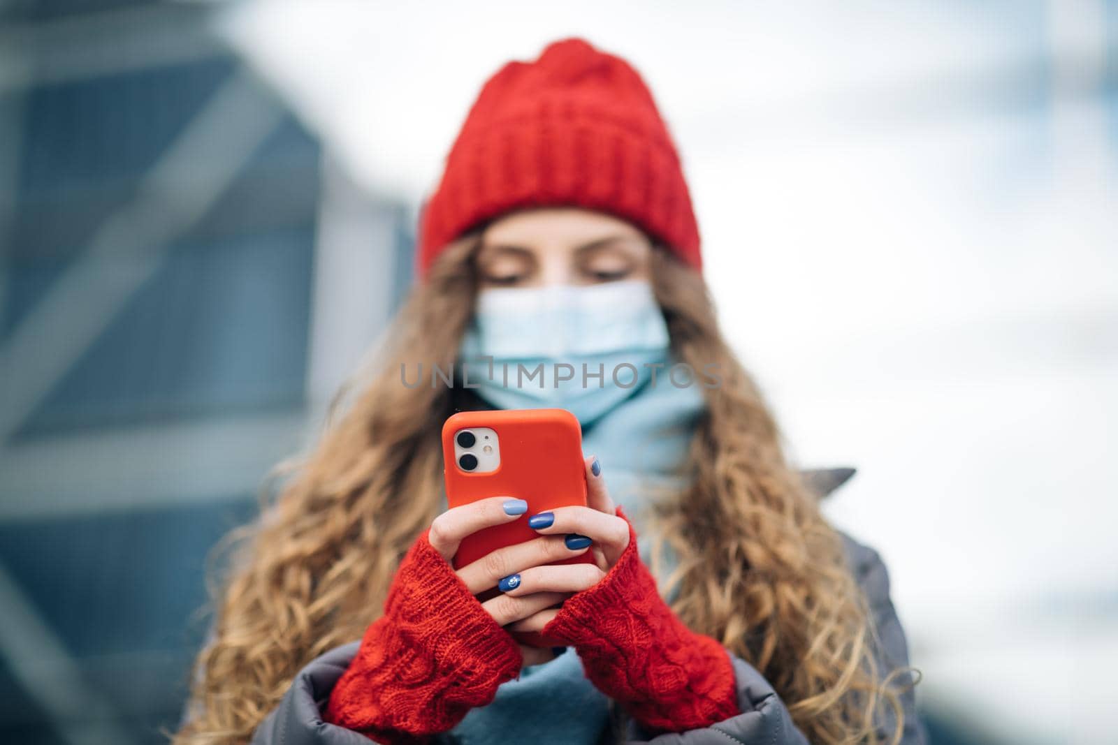Curly haired woman in protective medical mask walks down to the street uses phone texts scrolls surfs the internet search news covid19 coronavirus virus protection pandemic city