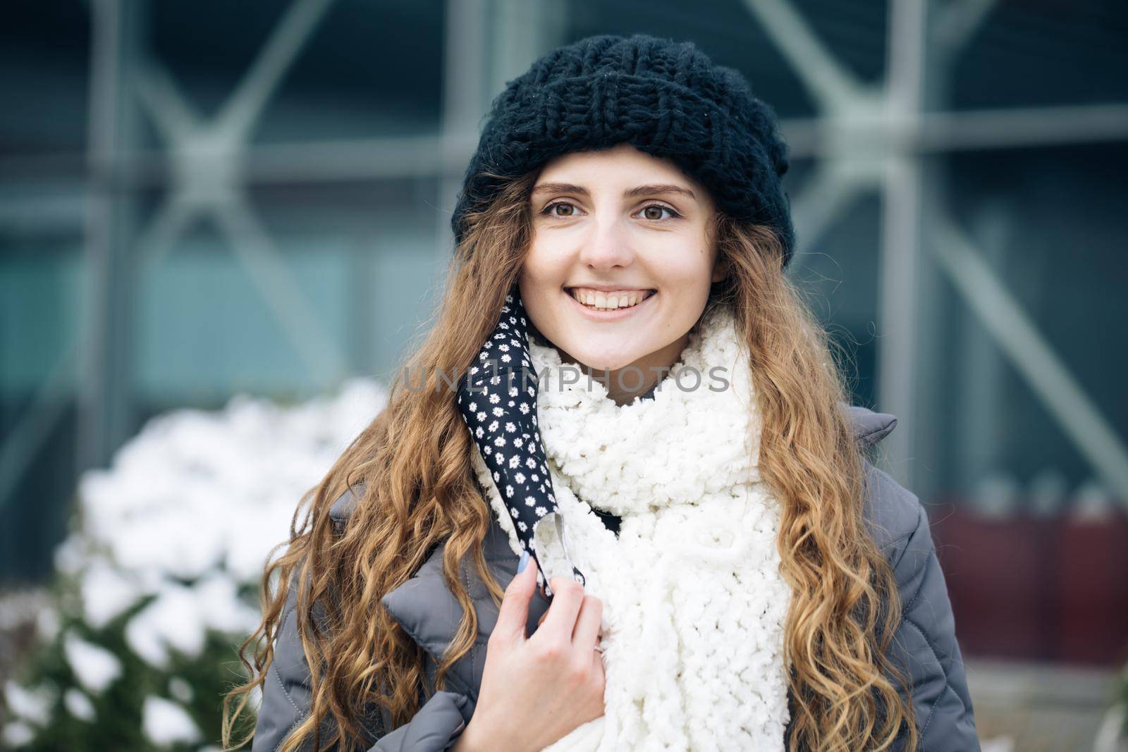 Woman takes off medical mask. Female breathes deeply and smiling looking at camera on city background. Health care and medical concept. Close up portrait curly female standing on winter street by uflypro