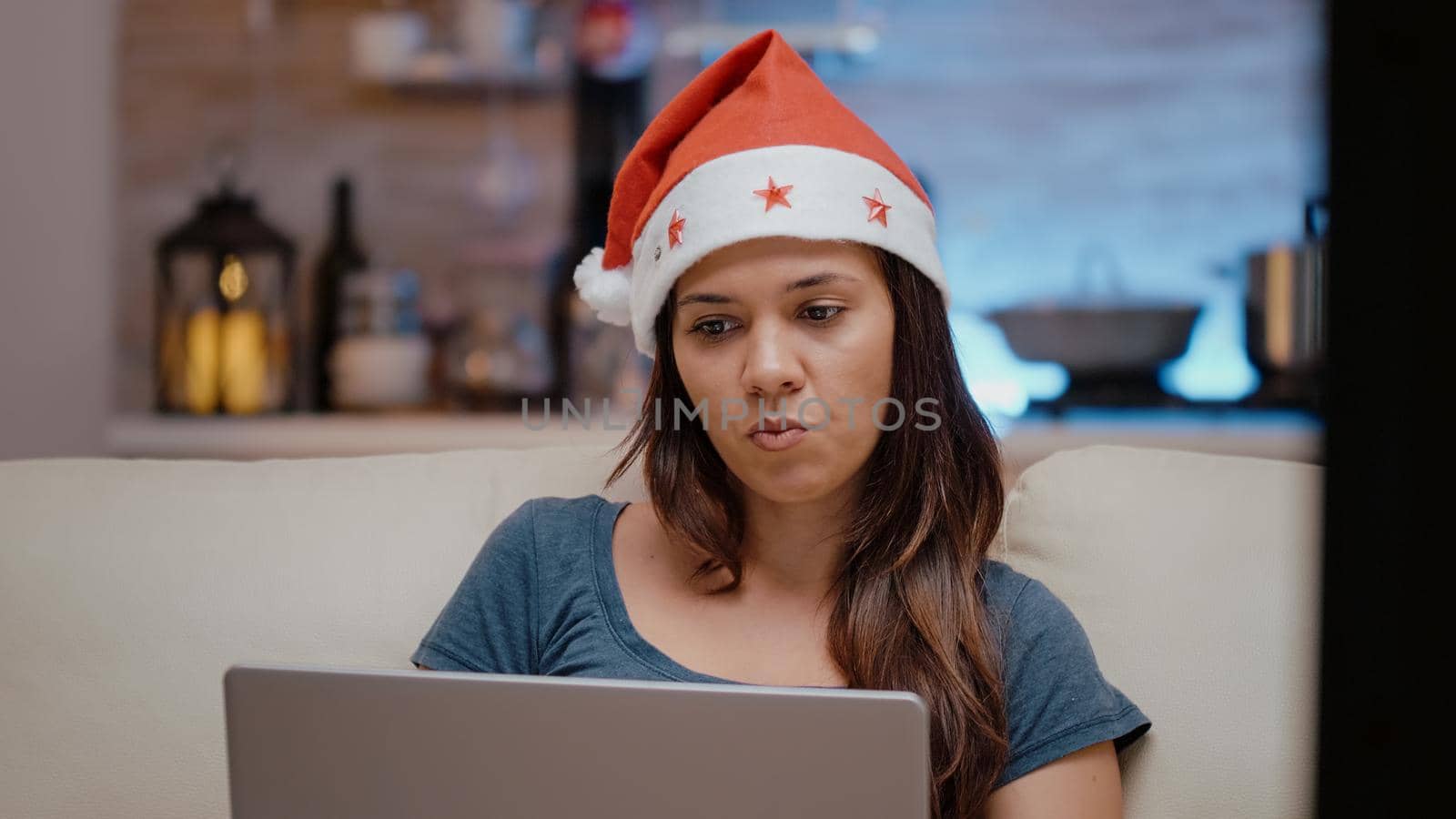 Festive person working with laptop and watching television on christmas holiday. Woman with santa hat looking at device and TV screen while eating chips on winter celebration