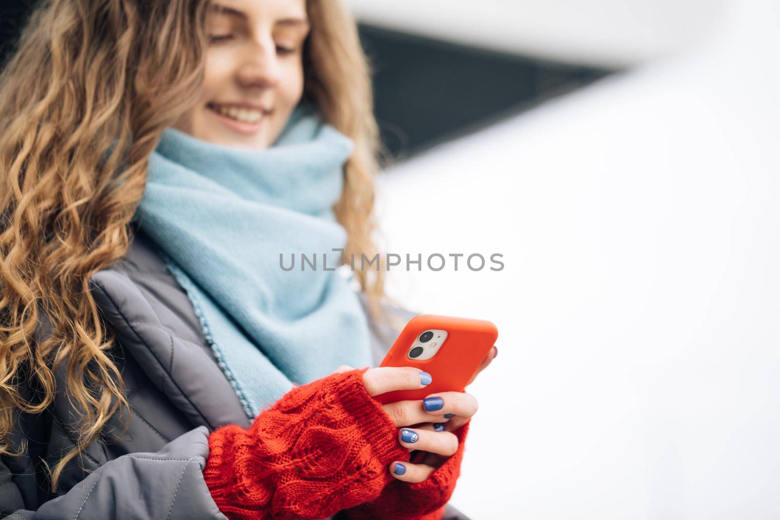 Portrait of cheerful Curly-haired Young female texting on smartphone standing on street in winter city on New Year. Female tapping on cellphone outdoors. Online shopping, buying new year's gifts