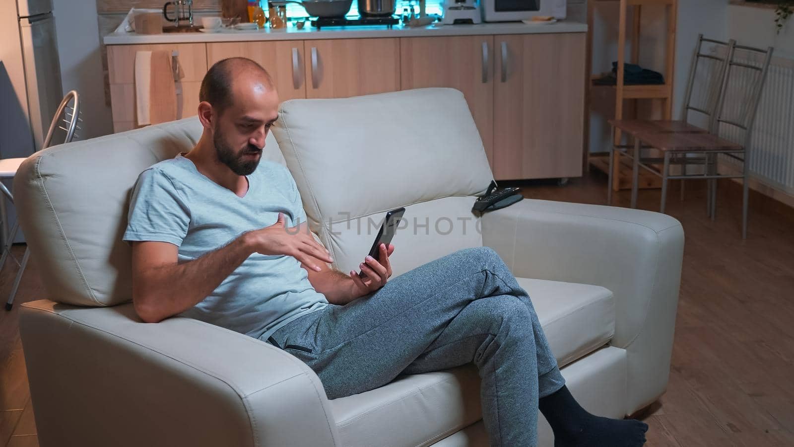 Focused adult talking with friends about social media connection using modern smartphone while sitting on couch. Tired man working late at night in kitchen using modern technology wireless