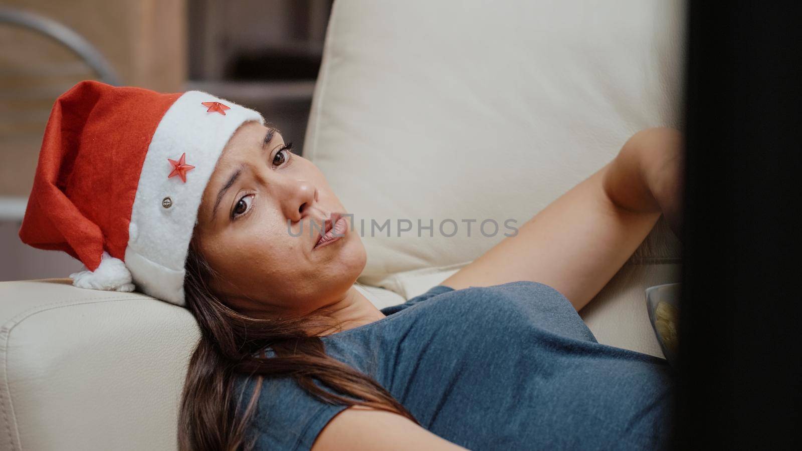 Close up of bored woman eating chips and watching TV while laying on couch. Person with santa hat looking at movie on television, celebrating christmas eve holiday alone at home