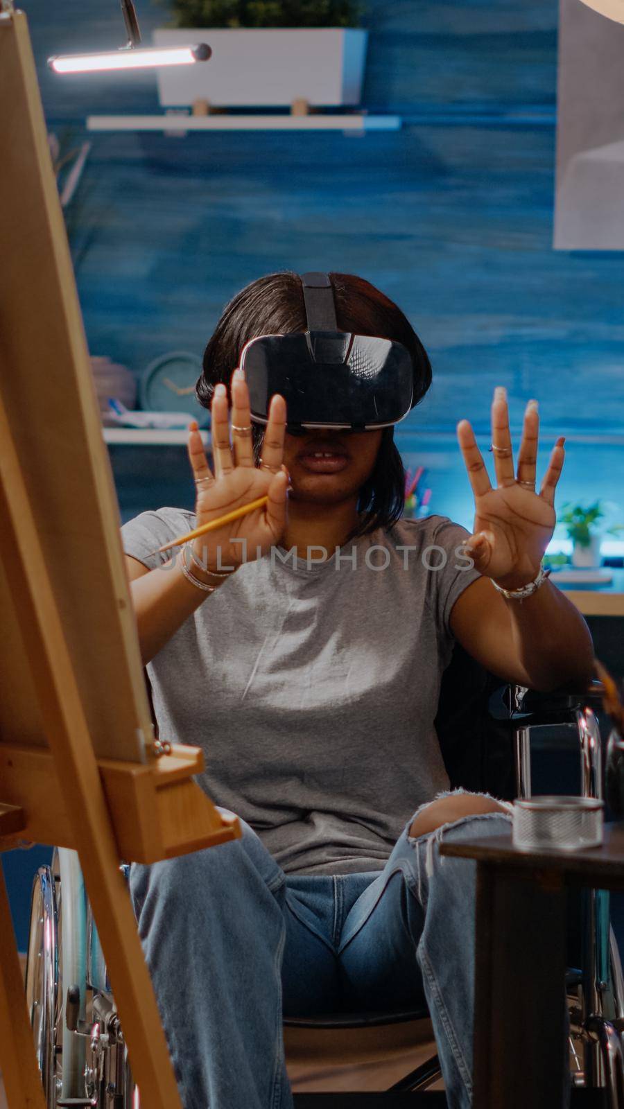 Black artist woman using vr glasses for art project in workshop room at home. African american person with handicap sitting in wheelchair while having headset for masterpiece