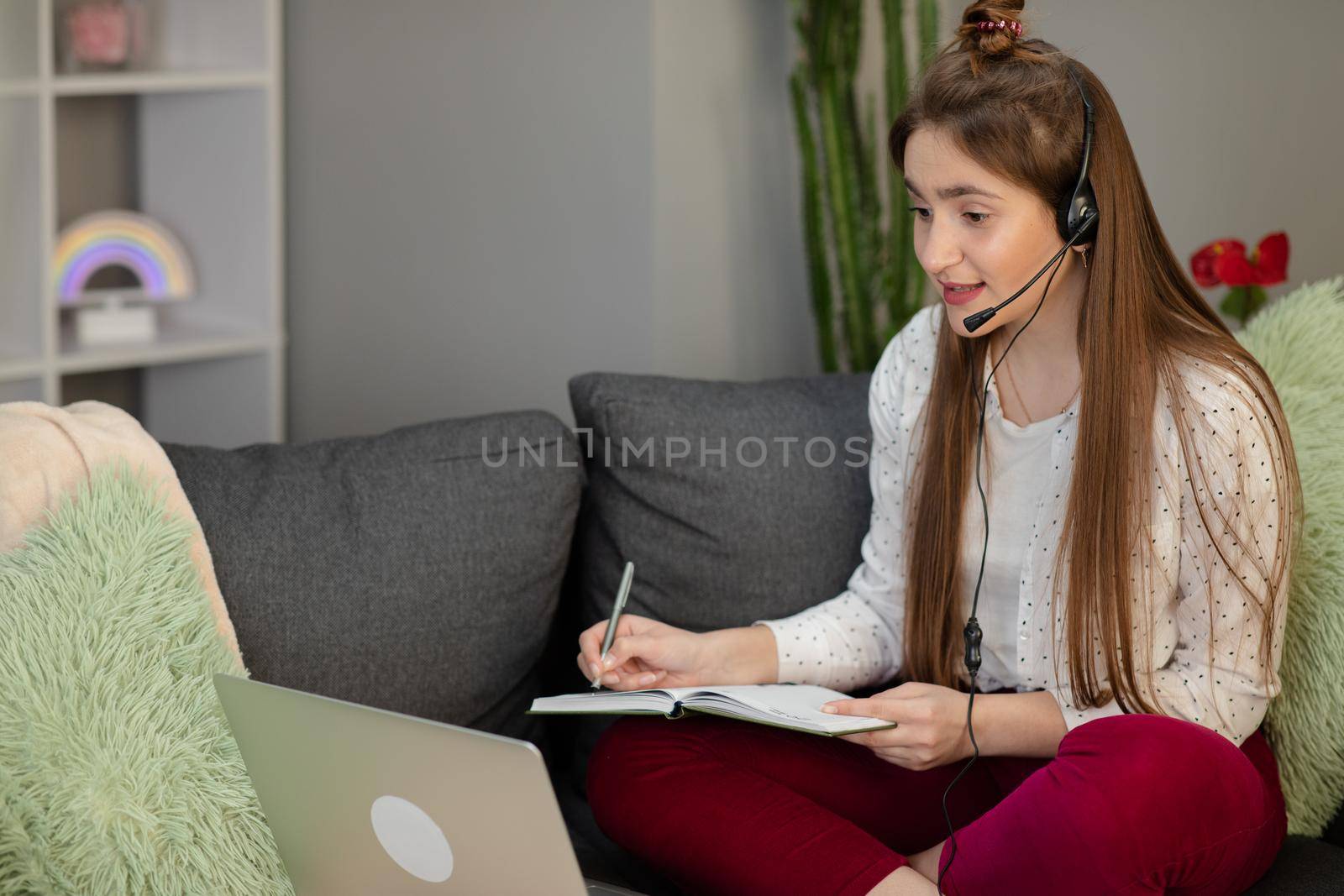 Smiling teenage girl wearing headphones using laptop sitting on bed. Happy teen school student conference calling on computer for online distance learning communicating with friend by webcam at home by uflypro