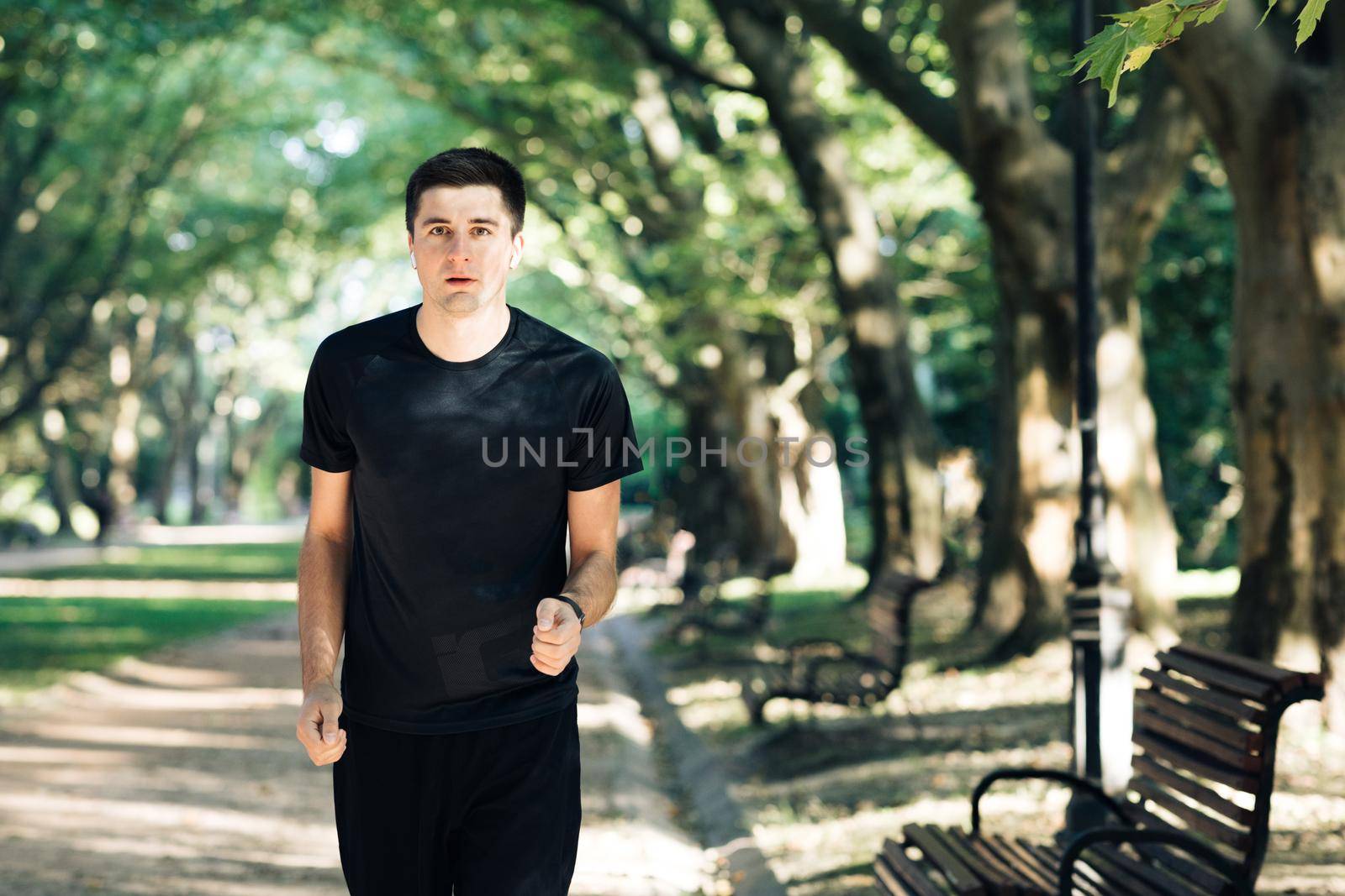 Adult man running in the city park using bluetooth earphones and smart watch app enjoying healthy active lifestyle. Male runner working out cardio exercise workout on summer setting sun. by uflypro