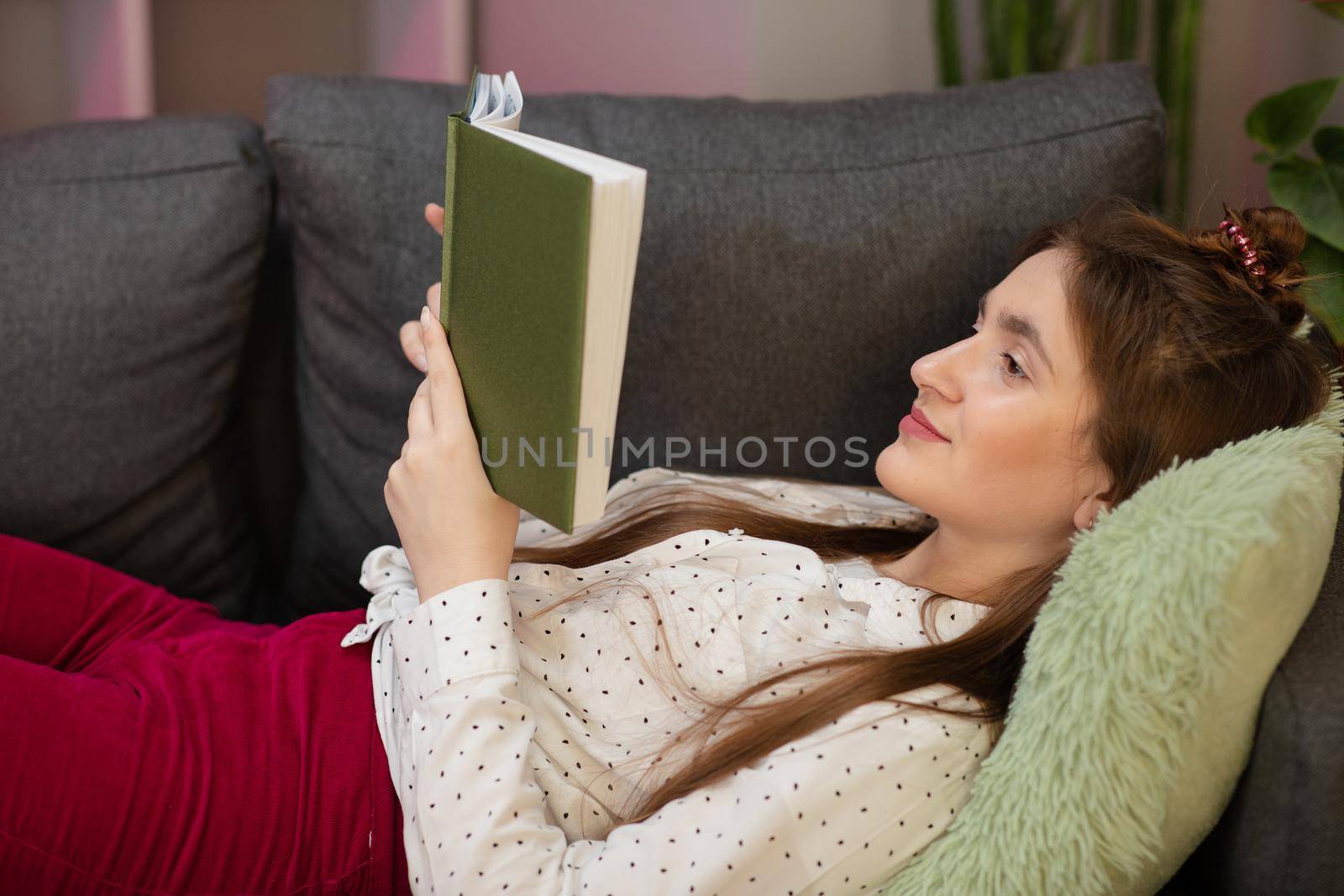 Portrait of smiling woman holding book and lying on couch. Teenager girl home - student read book laying on sofa