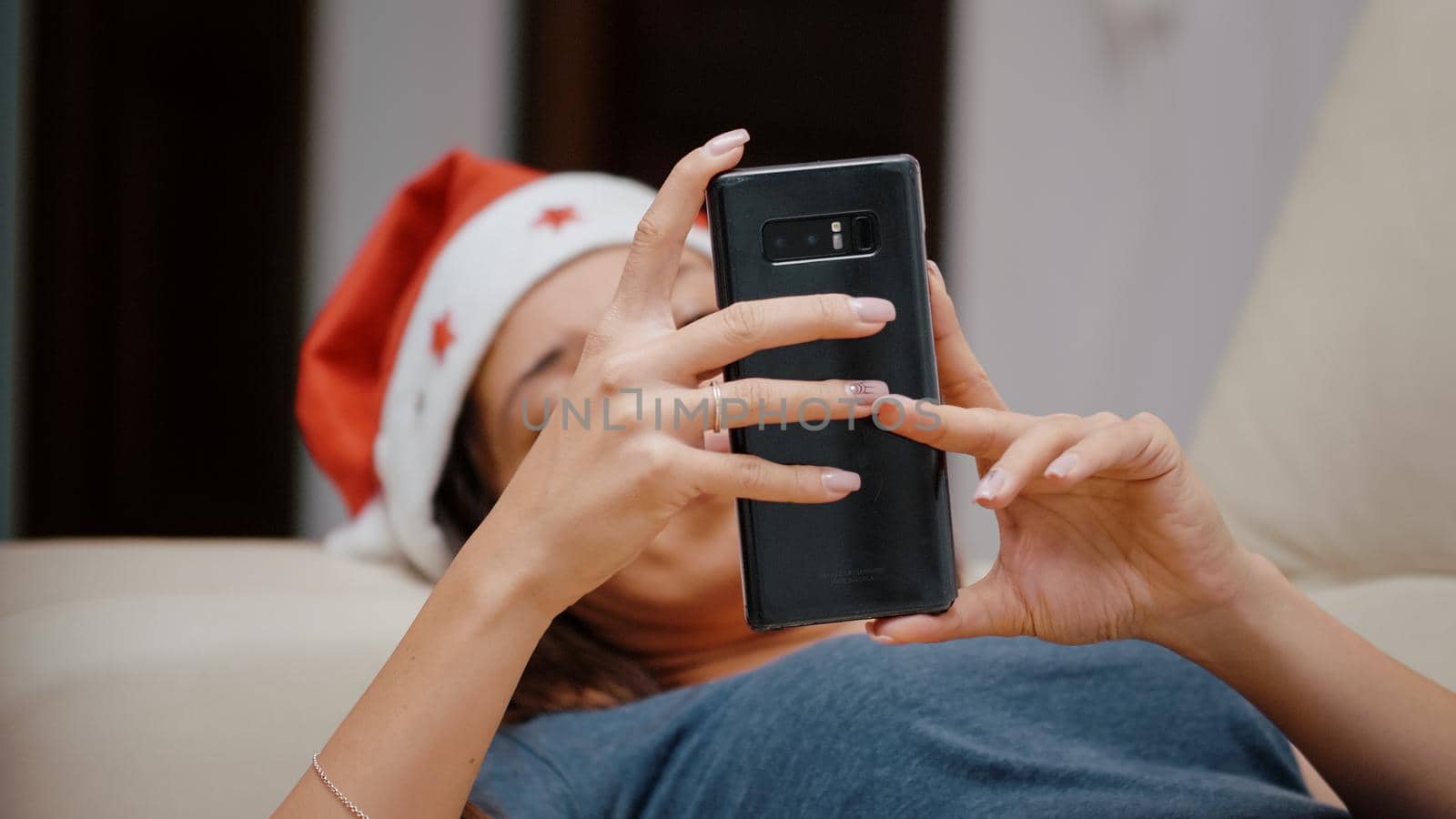 Close up of festive woman looking at smartphone and TV for christmas eve celebration. Person holding gadget and watching television while laying on couch on winter holiday. Adult with device