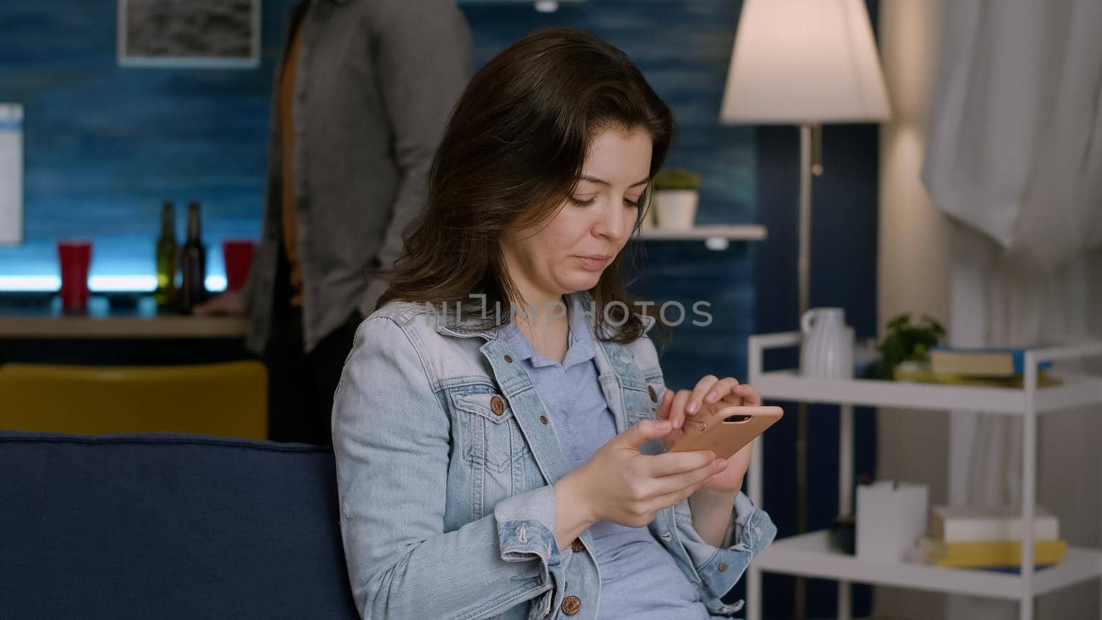 Cheerful woman browsing on social media using smartphone while resting on couch late at night in living room. In background her friends drinking beer, having fun during wekeend party