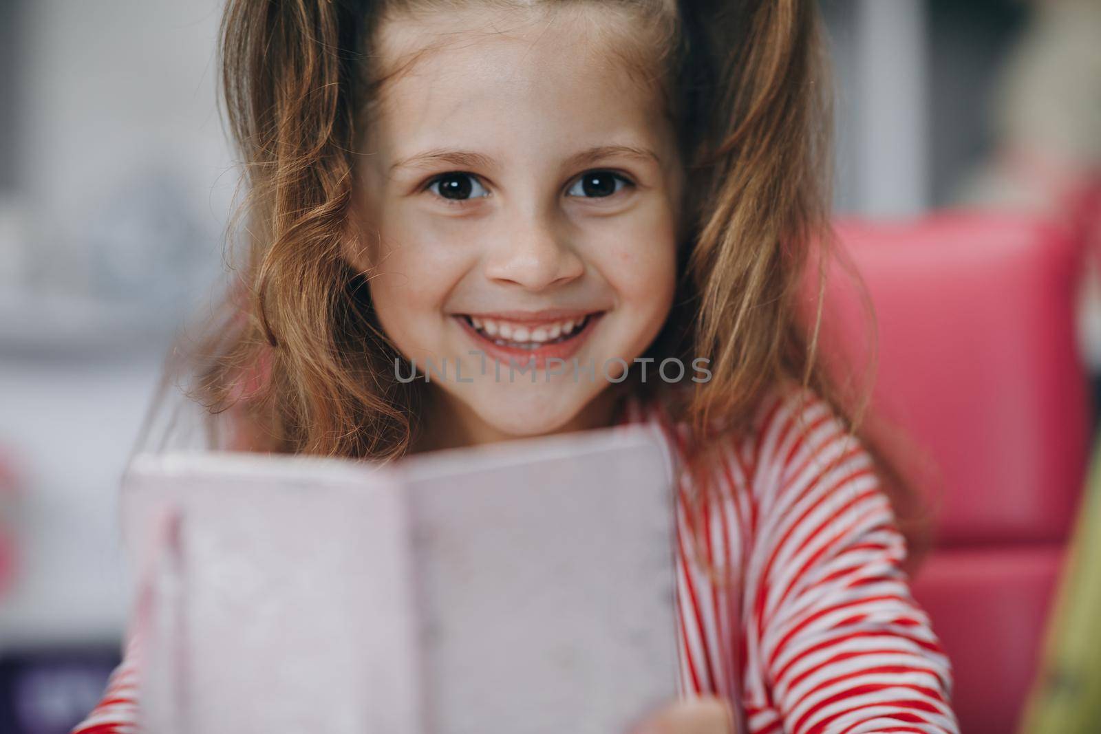 Portrait of a preschool girl looking at the camera and smiling. Kid girl sitting at the desk in children room at home and studying online. Distance learning, school online.