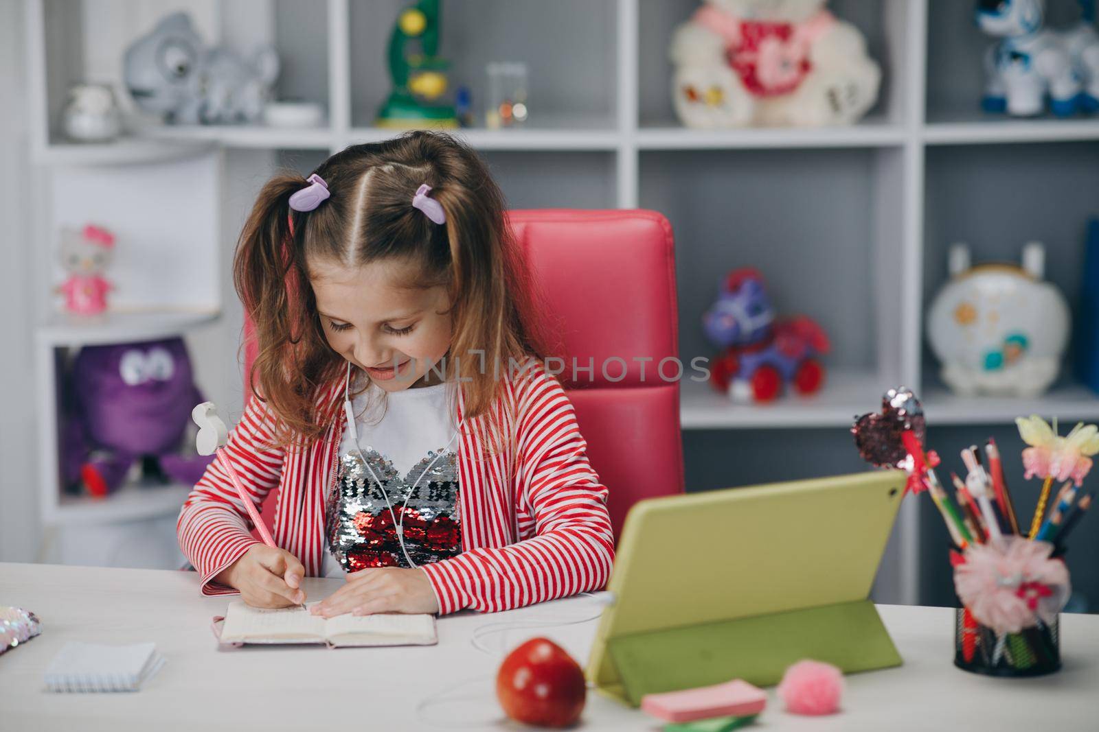 Preschool girl watching lesson online and studying from home. Kid girl taking notes while looking at computer screen by uflypro