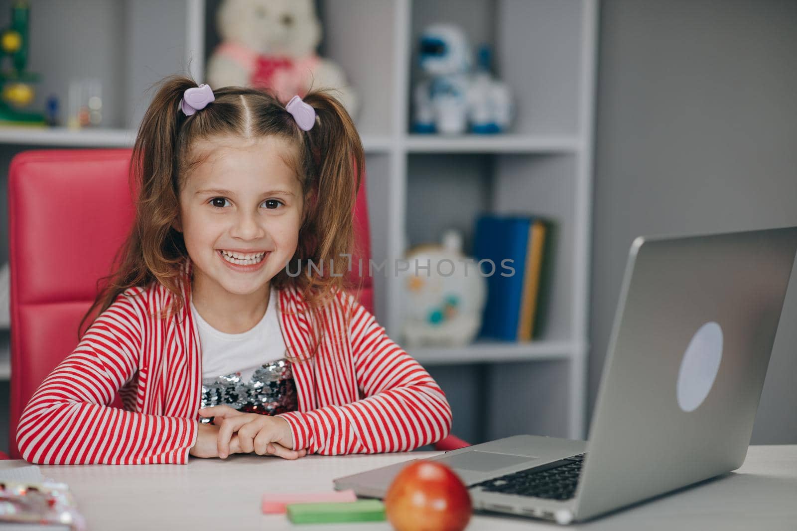 Portrait of a preschool girl looking at the camera and smiling. Kid girl sitting at the desk in children room at home and studying online. Distance learning, school online by uflypro