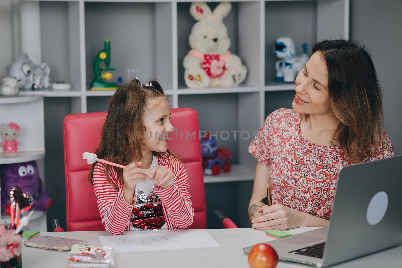 Woman tutor or foster parent mum helping cute caucasian school child girl doing homework sitting at kitchen table. Diverse nanny and kid learning writing in notebook studying at home.