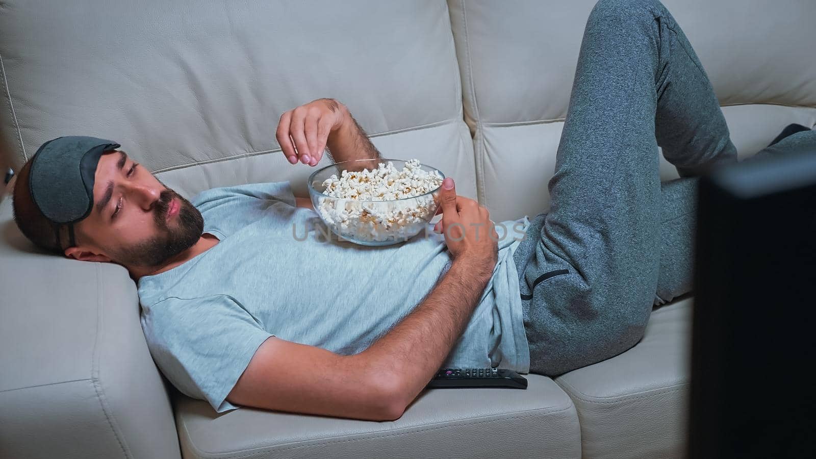 Tired man relaxing on sofa in front of television eating popcorn while watching movie show by DCStudio