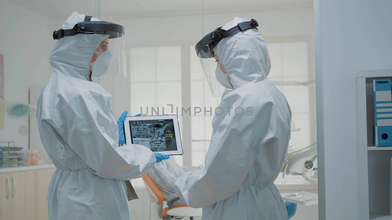 Team of dentists wearing ppe suits while looking at x ray teeth scan on digital tablet for orthodontic consultation. Caucasian stomatologists examining dental patient radiography