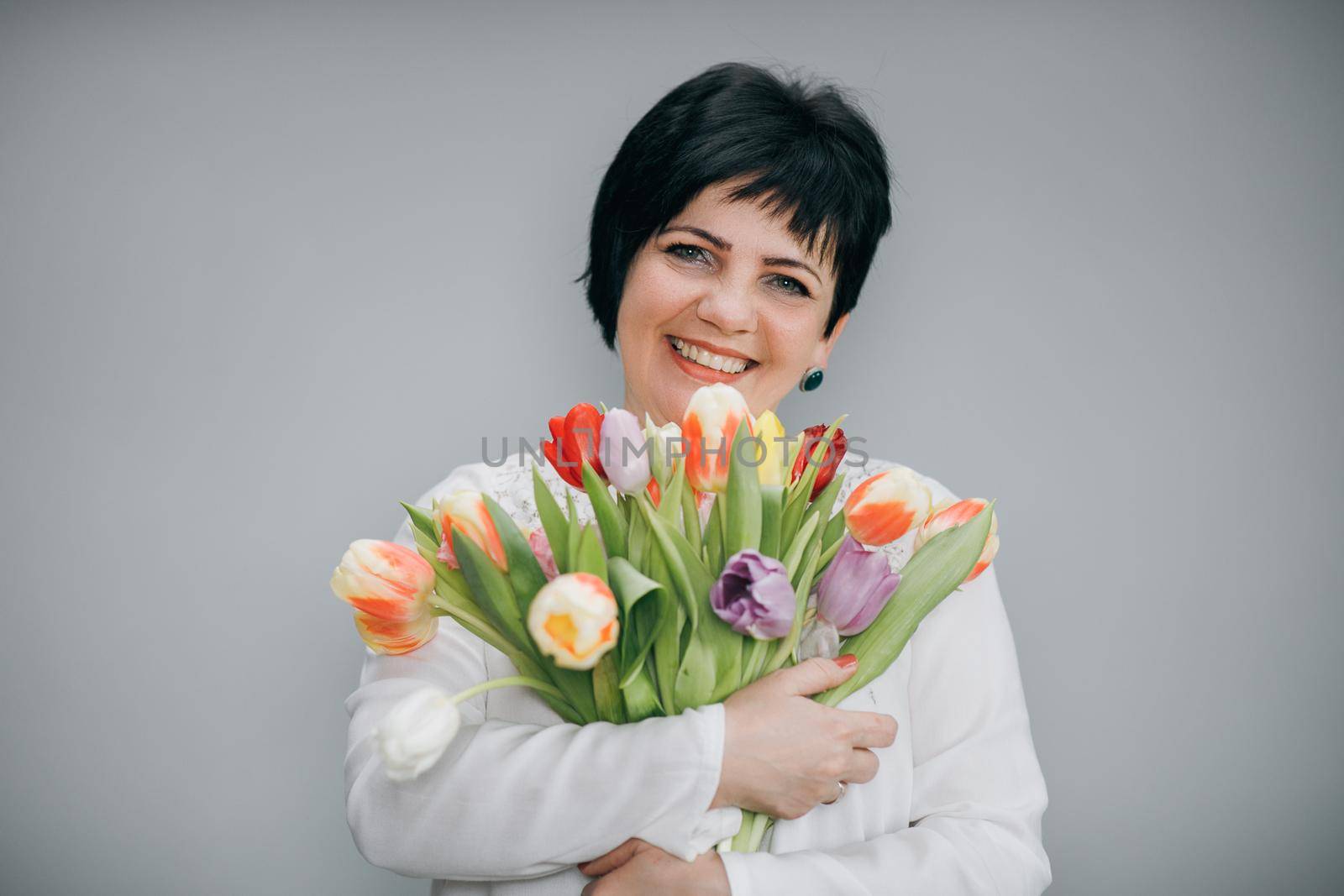 Closeup portrait of elderly woman holding flower bouquet in her hands. Model posing isolated over grey studio background. Charming female 8 march concept.