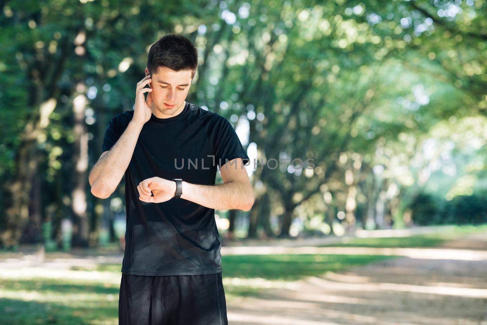 Man in summer sports uniform running in the city park with smartwatch for measuring speed, distance and heart rate. Wireless earphones. Training outside.