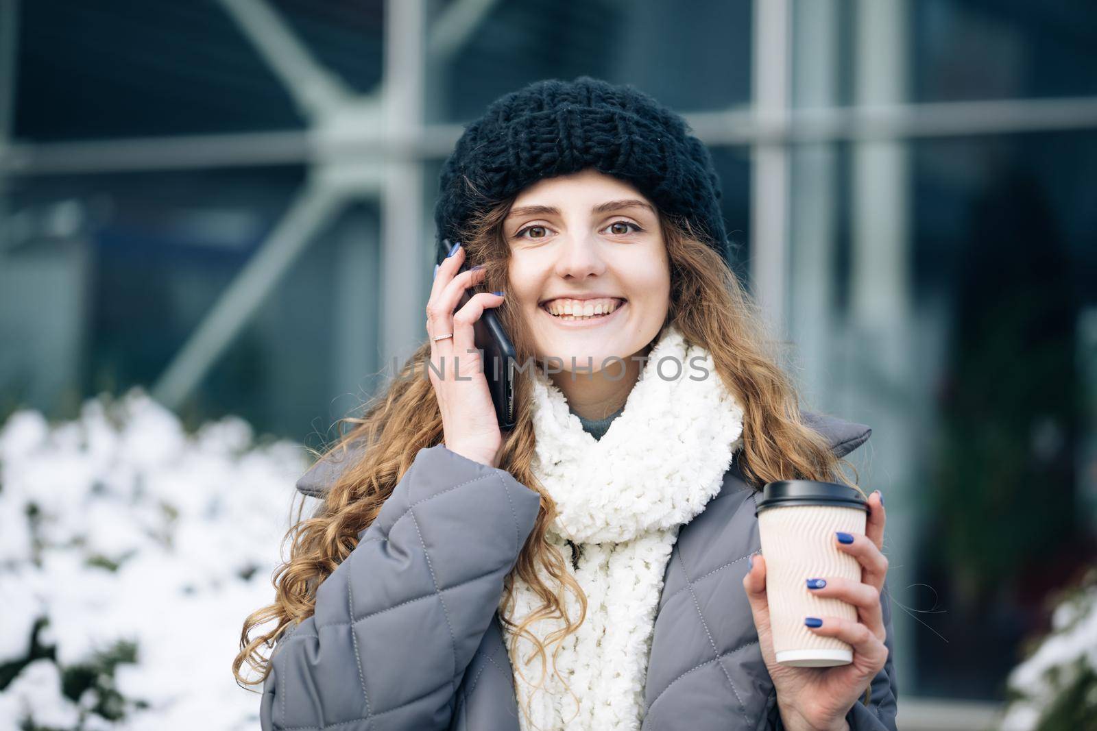 Curly haired woman using her smartphone and holding paper cup with coffee. Young girl talking on her mobile phone while walking in the city street by uflypro