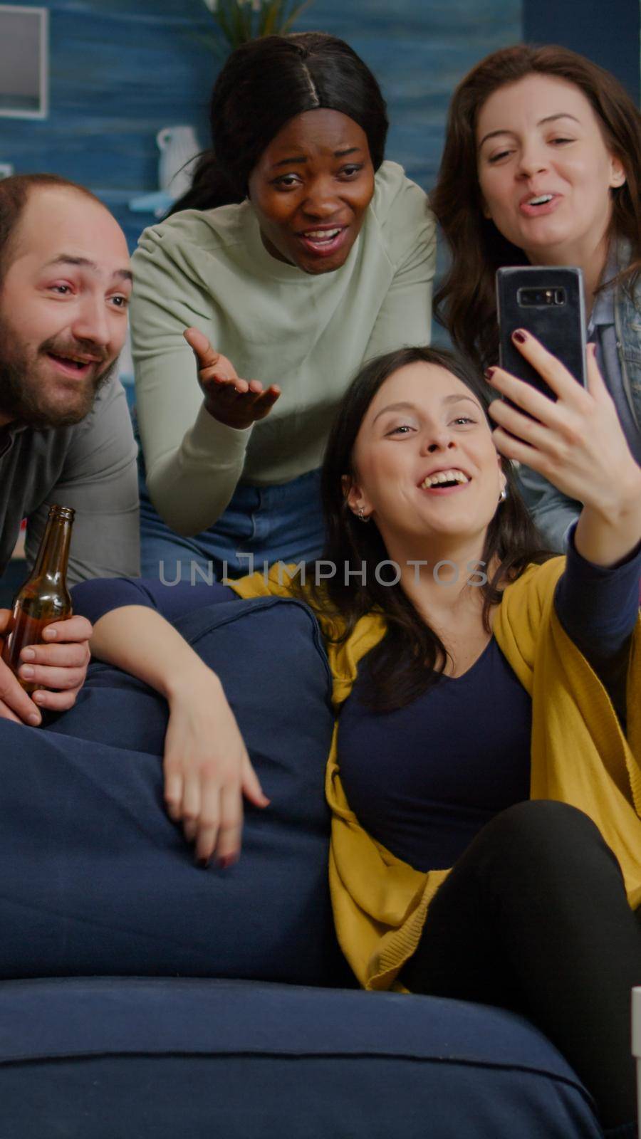 Multiracial friends speaking with collegue man during video call conference using modern smartphone. Group of multi-ethnic people hanging out together, drinking beer, having fun during night party
